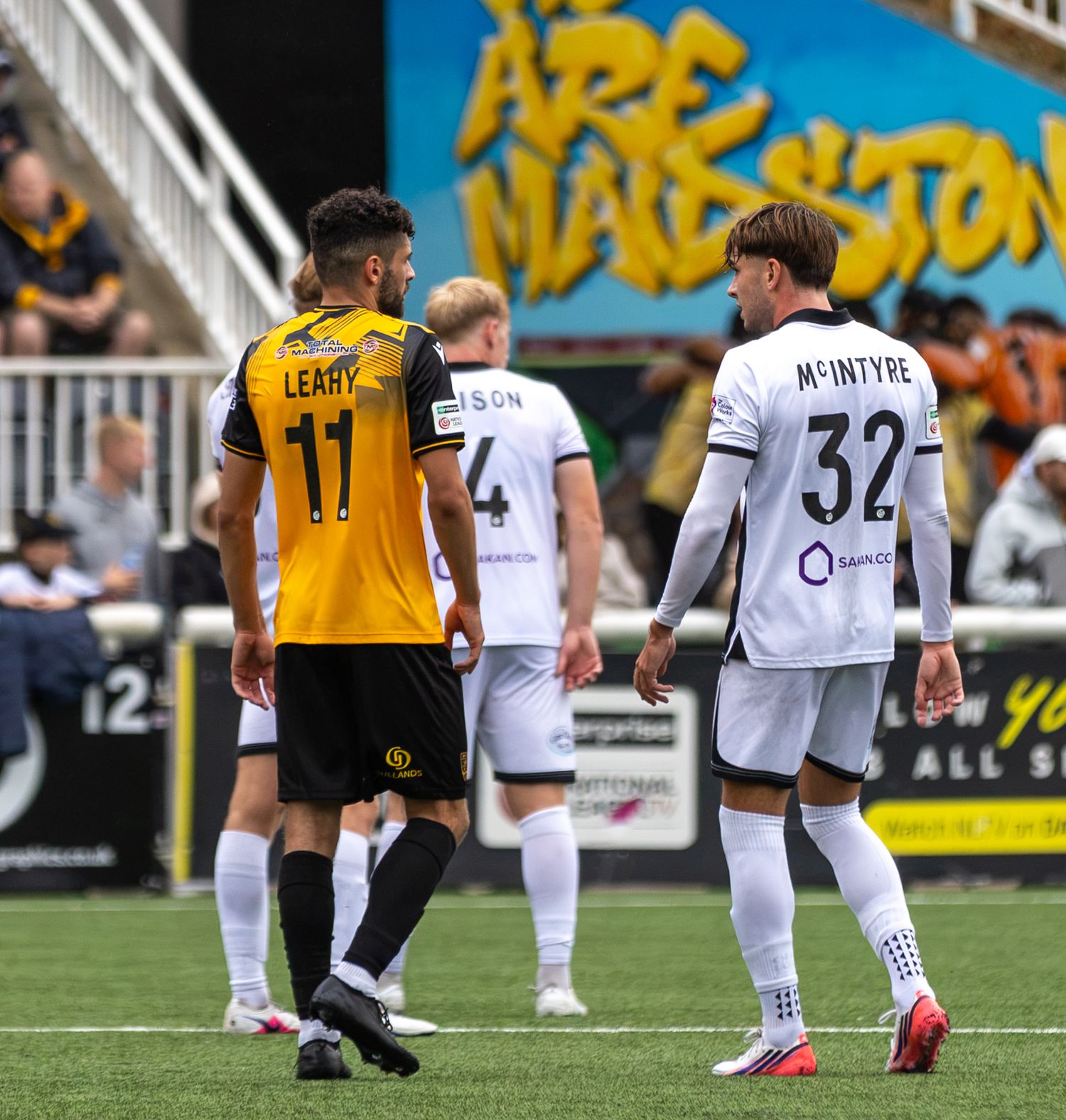 Football players LEARY 11 and MCINTYRE 32 face off on the pitch during a competitive match at a packed stadium.