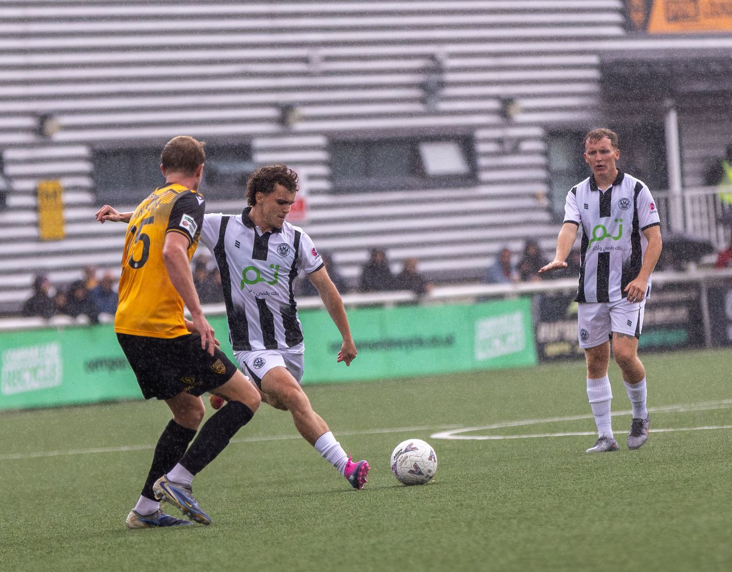 Soccer players in black and white striped kits compete for the ball during a match at a green stadium.