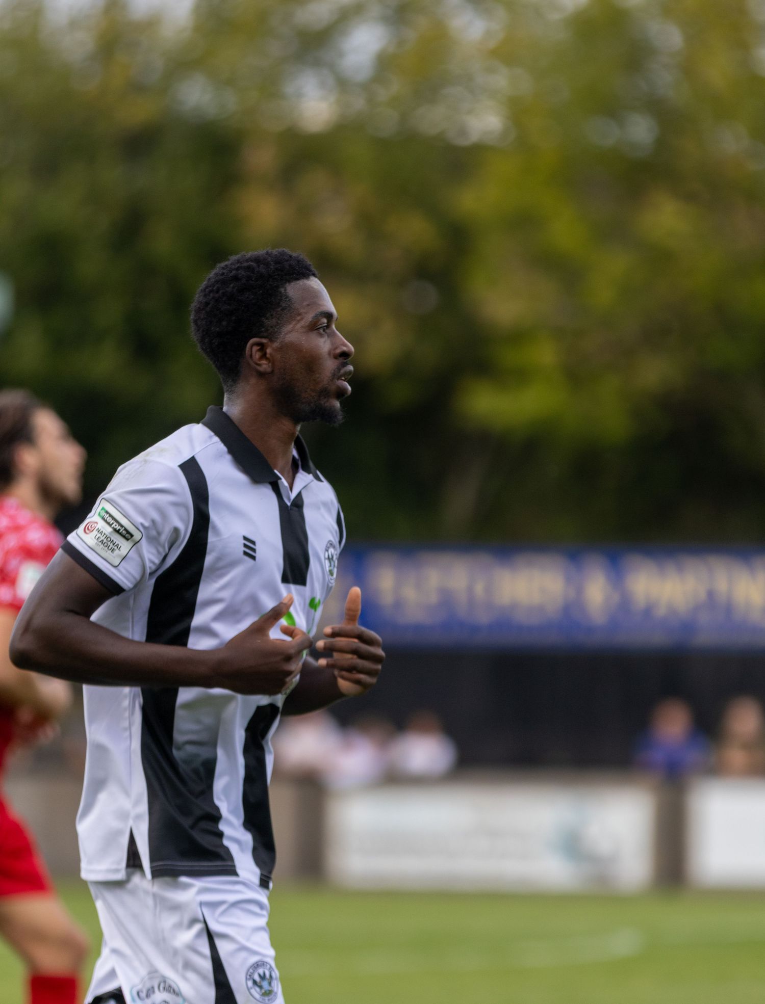 A footballer in black and white striped kit jogs on a grass pitch during an outdoor match.