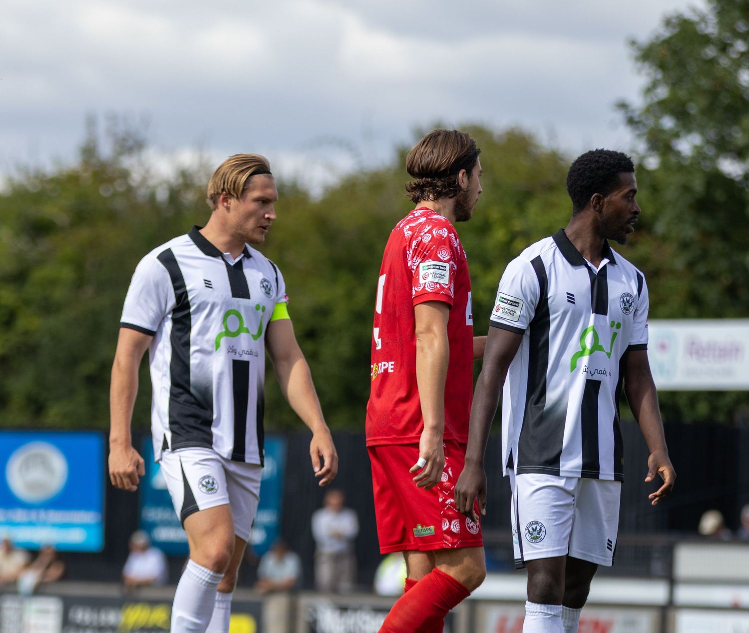 Three football players, two in black-white and one in red, walk across the pitch during a match.