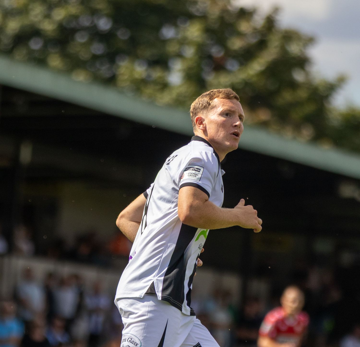 Blonde footballer in white and grey kit sprints across green pitch during sunny football match.