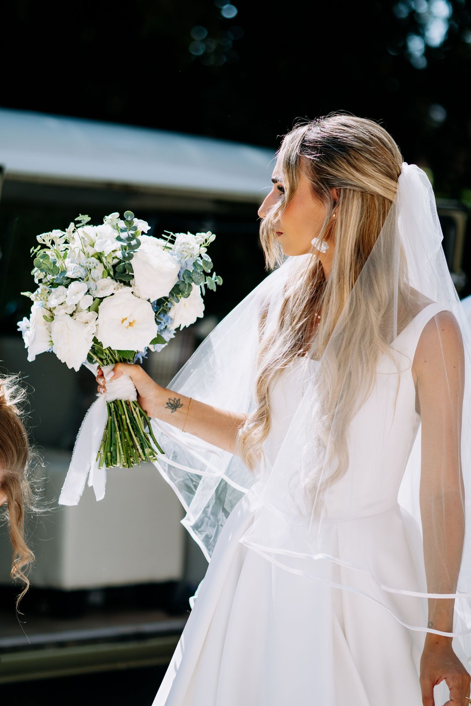 Side profile of bride in white gown and veil holding white bouquet near vintage vehicle.