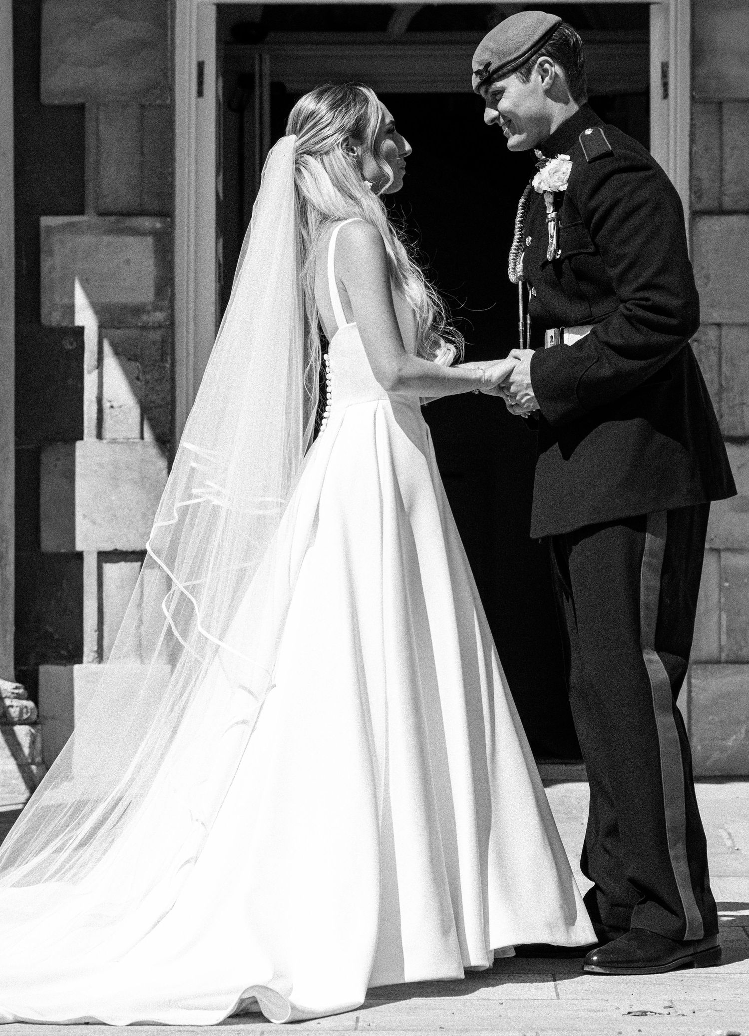 Black and white photo of bride in flowing veil and gown holding hands with groom in military uniform.