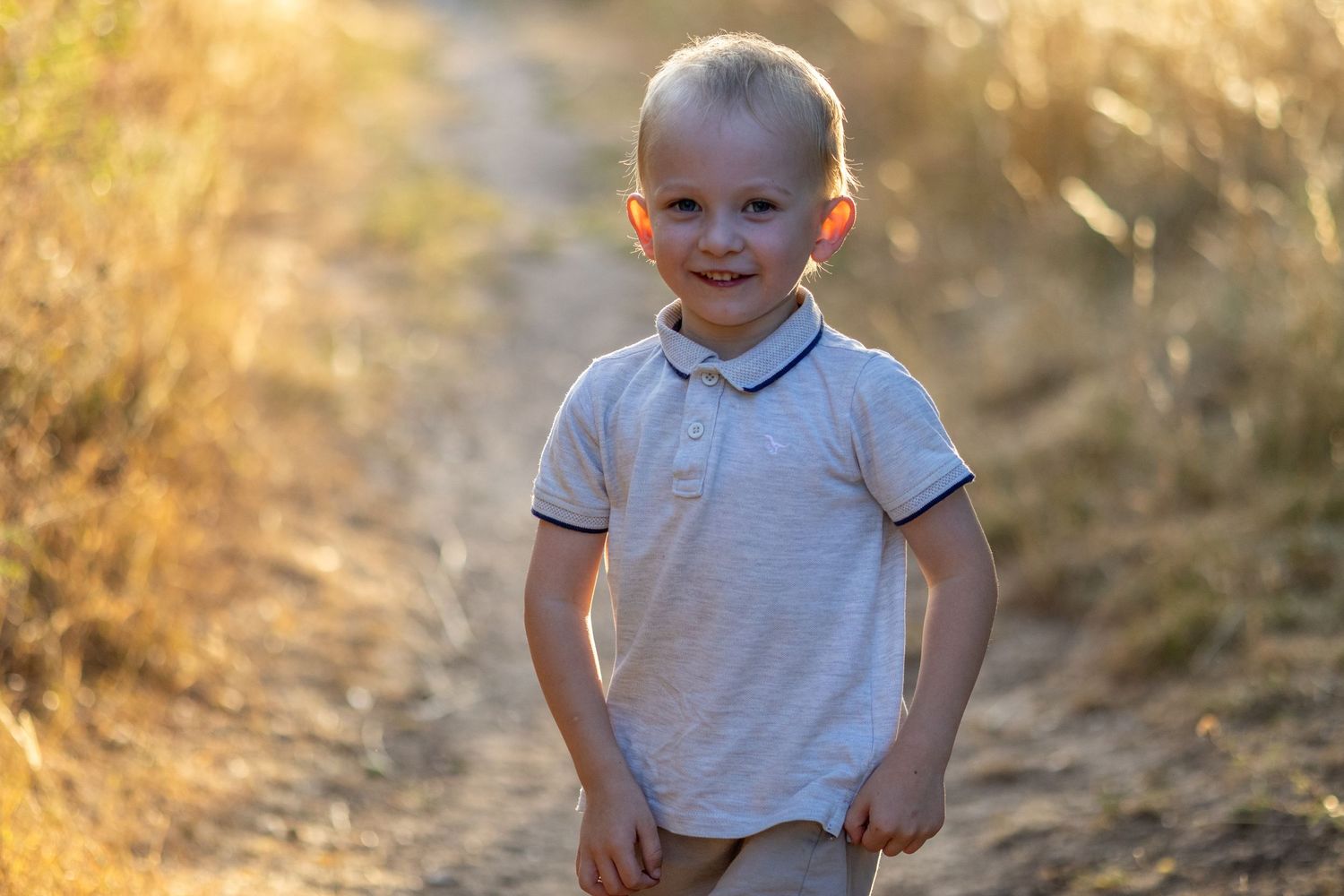A cheerful blonde toddler boy in a white polo shirt smiling on a sunny dirt path.