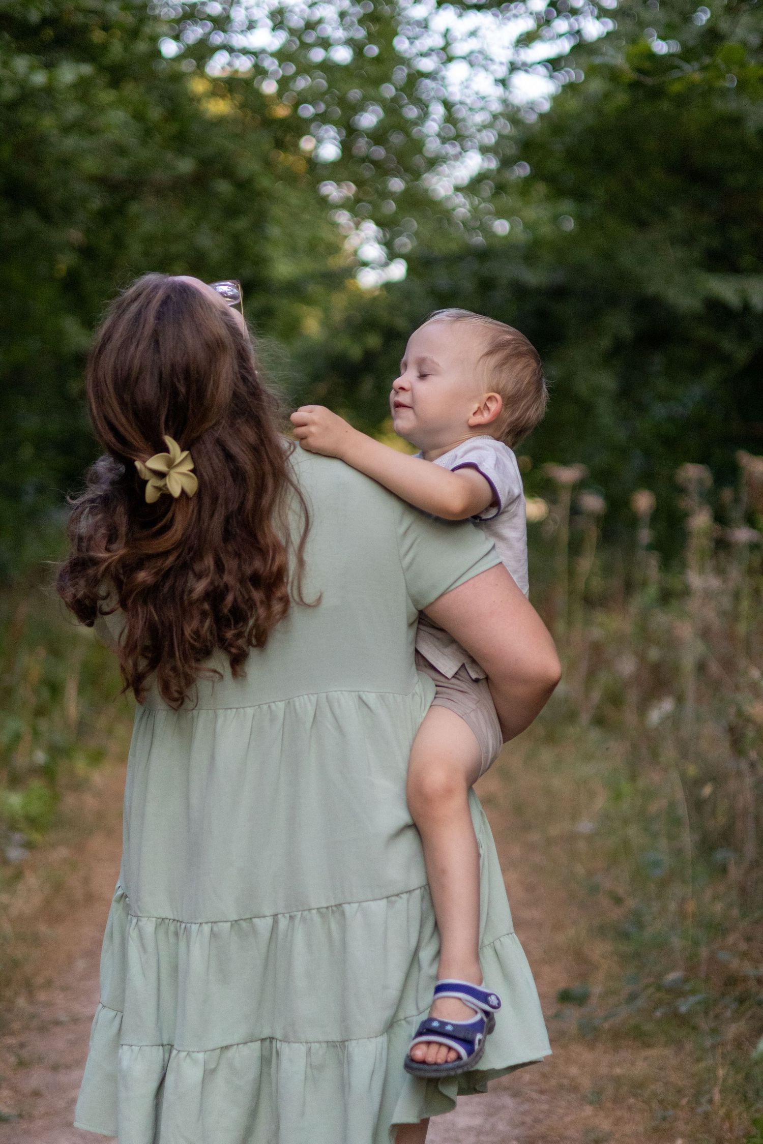 Mother in sage green dress holding smiling baby on a nature path surrounded by trees.