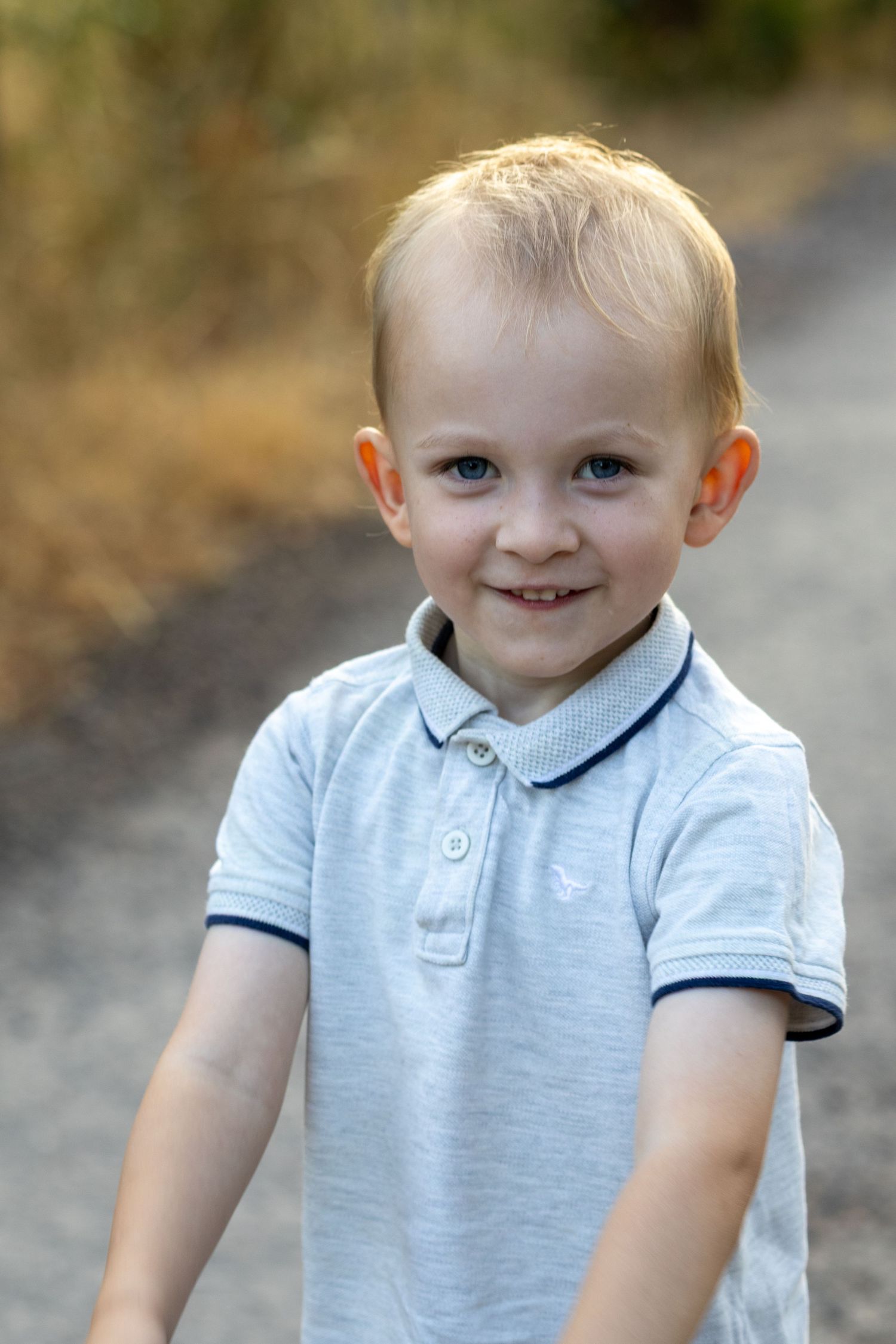 A cute blonde toddler in a grey polo shirt smiles brightly on an outdoor dirt path.