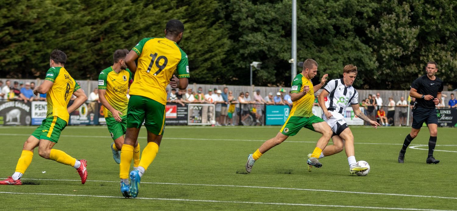 White-kitted player drives forward with the ball against yellow-green defenders during a sunny non-league football match.