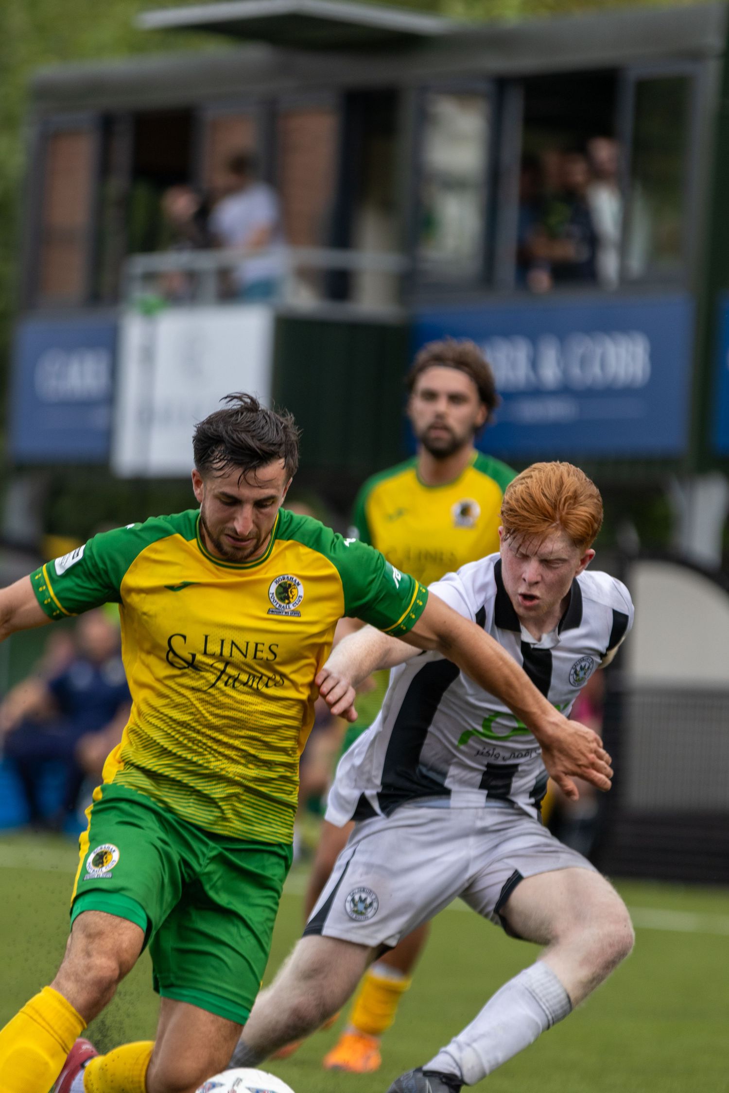 Two soccer players battle for the ball during a match, one in yellow-green and one in white-black kit.