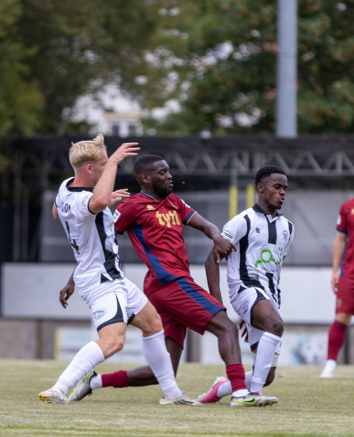 White-kitted players compete against maroon opponent in physical midfield battle during football match.