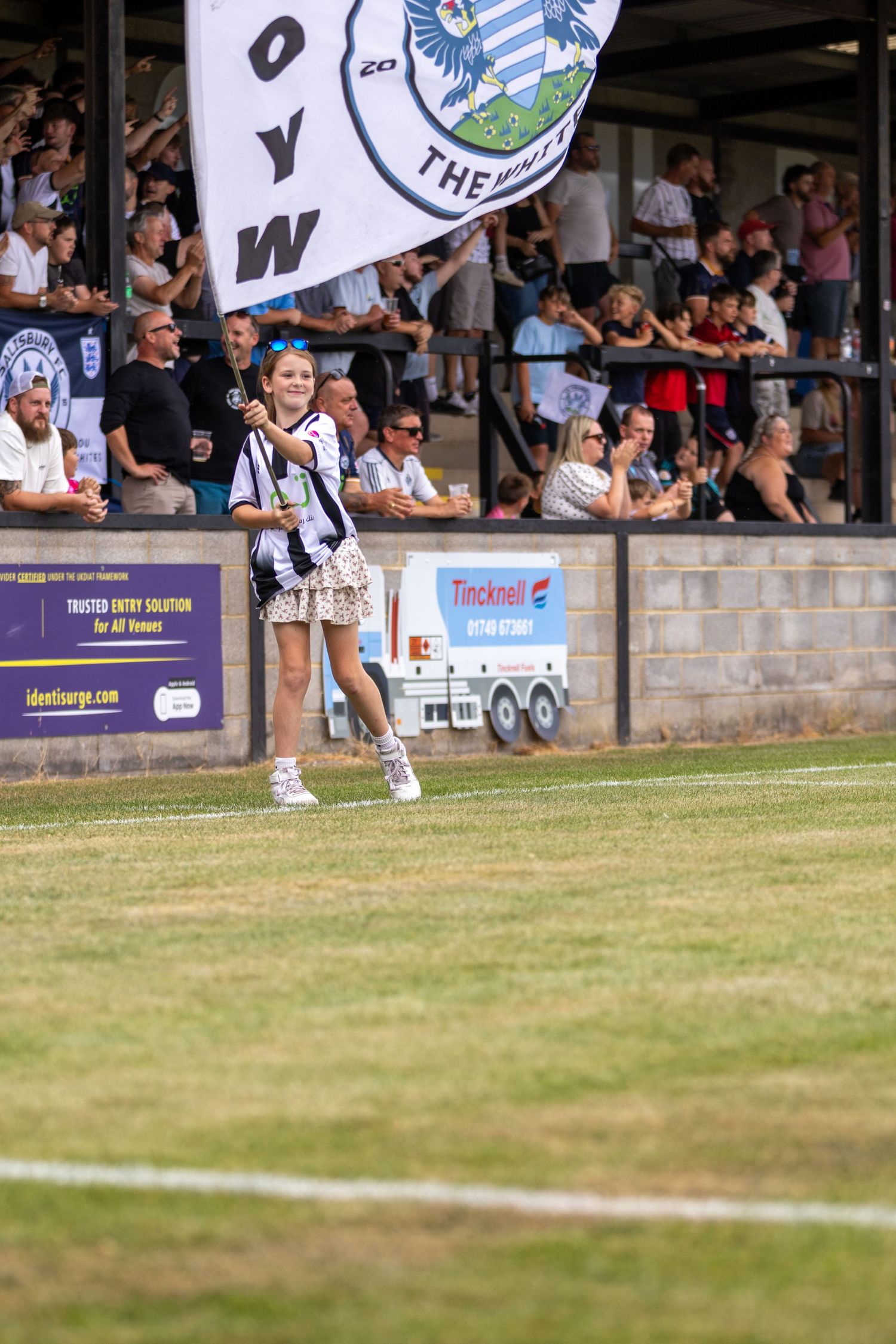 A young girl carries a large Salisbury FC flag in front of a packed stadium stand on a sunny matchday.