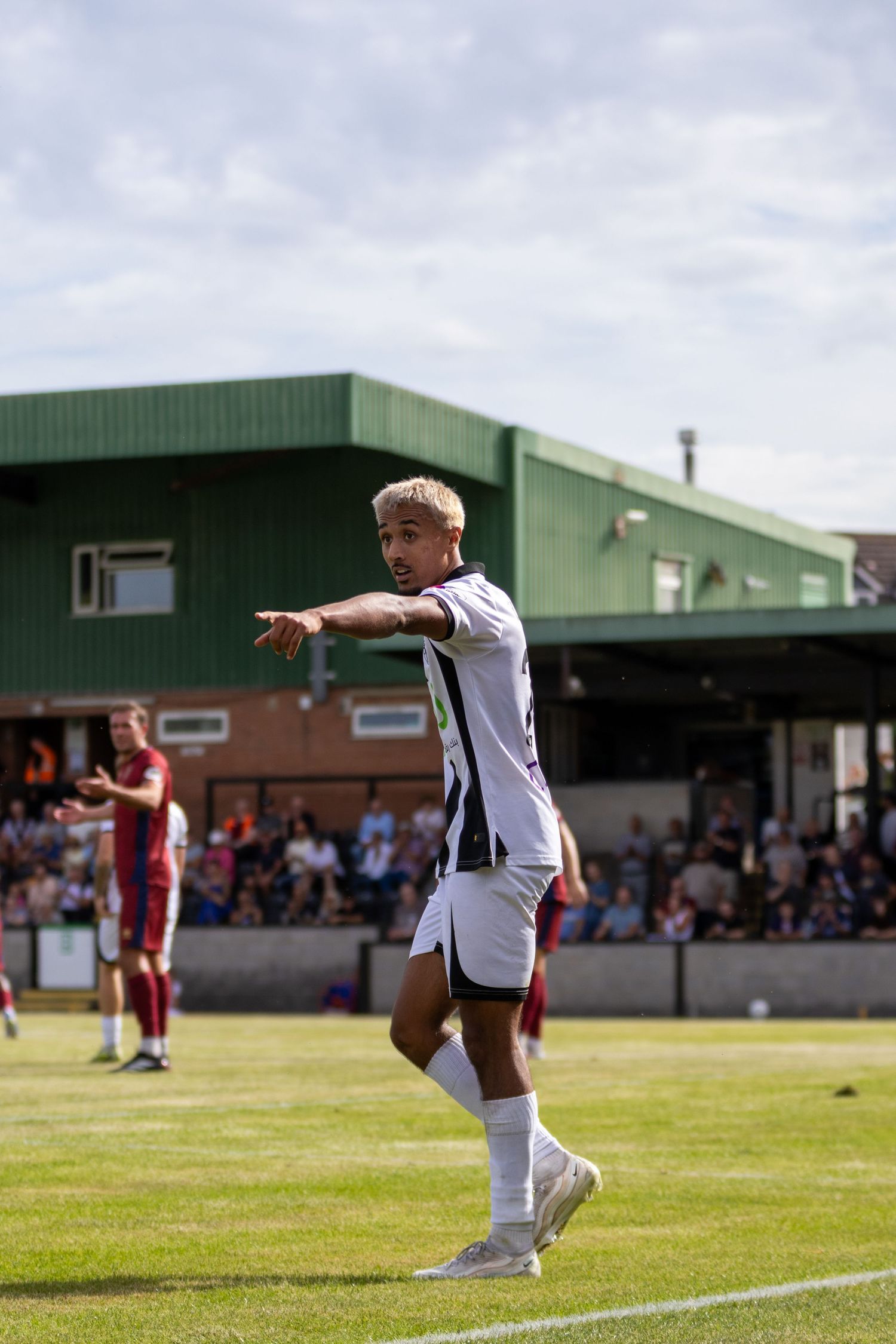 A footballer in black and white kit points directions during a sunny non-league match with spectators in background.