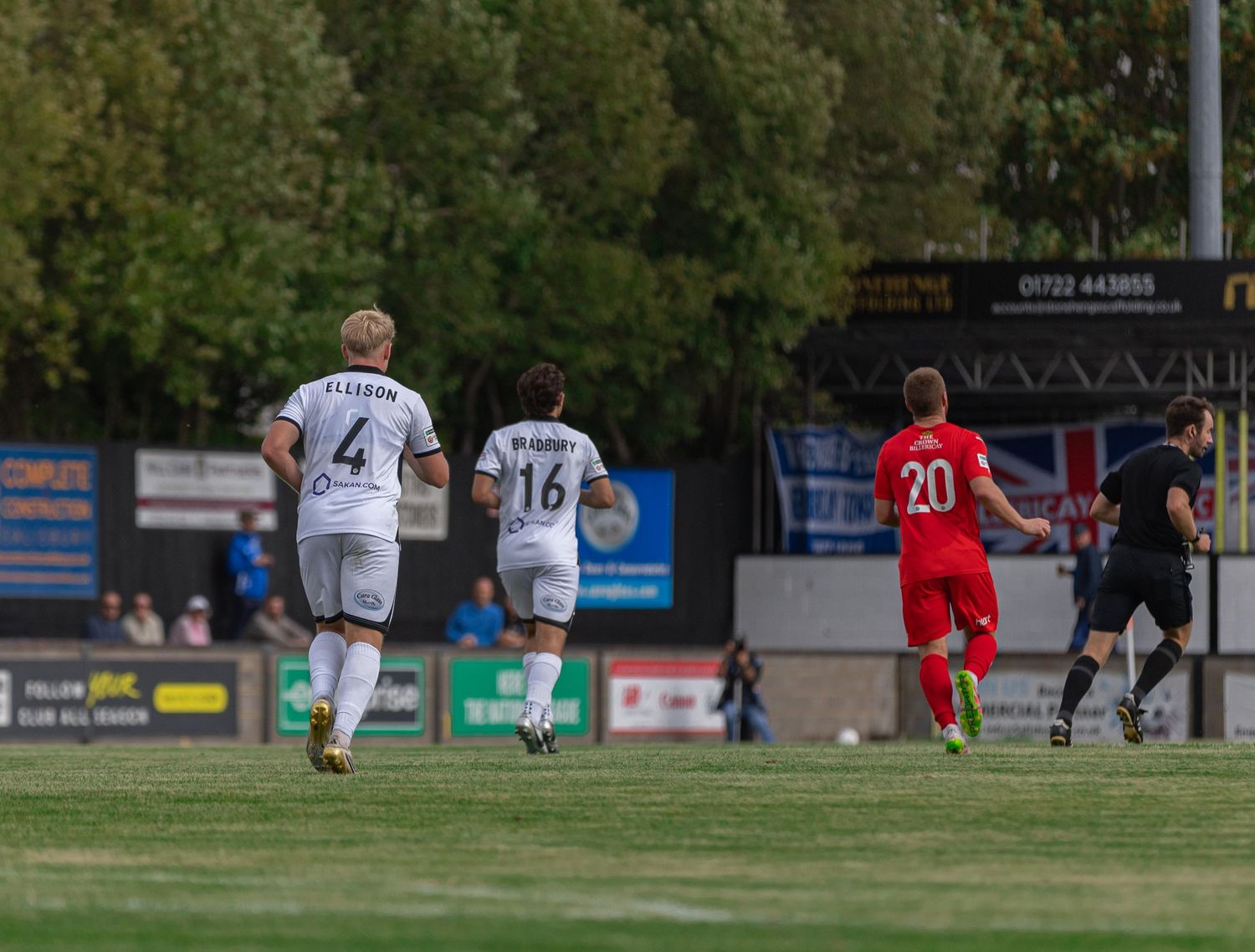 Soccer players in white jerseys numbered 4 and 16 run alongside a player in red number 20 during an outdoor match.