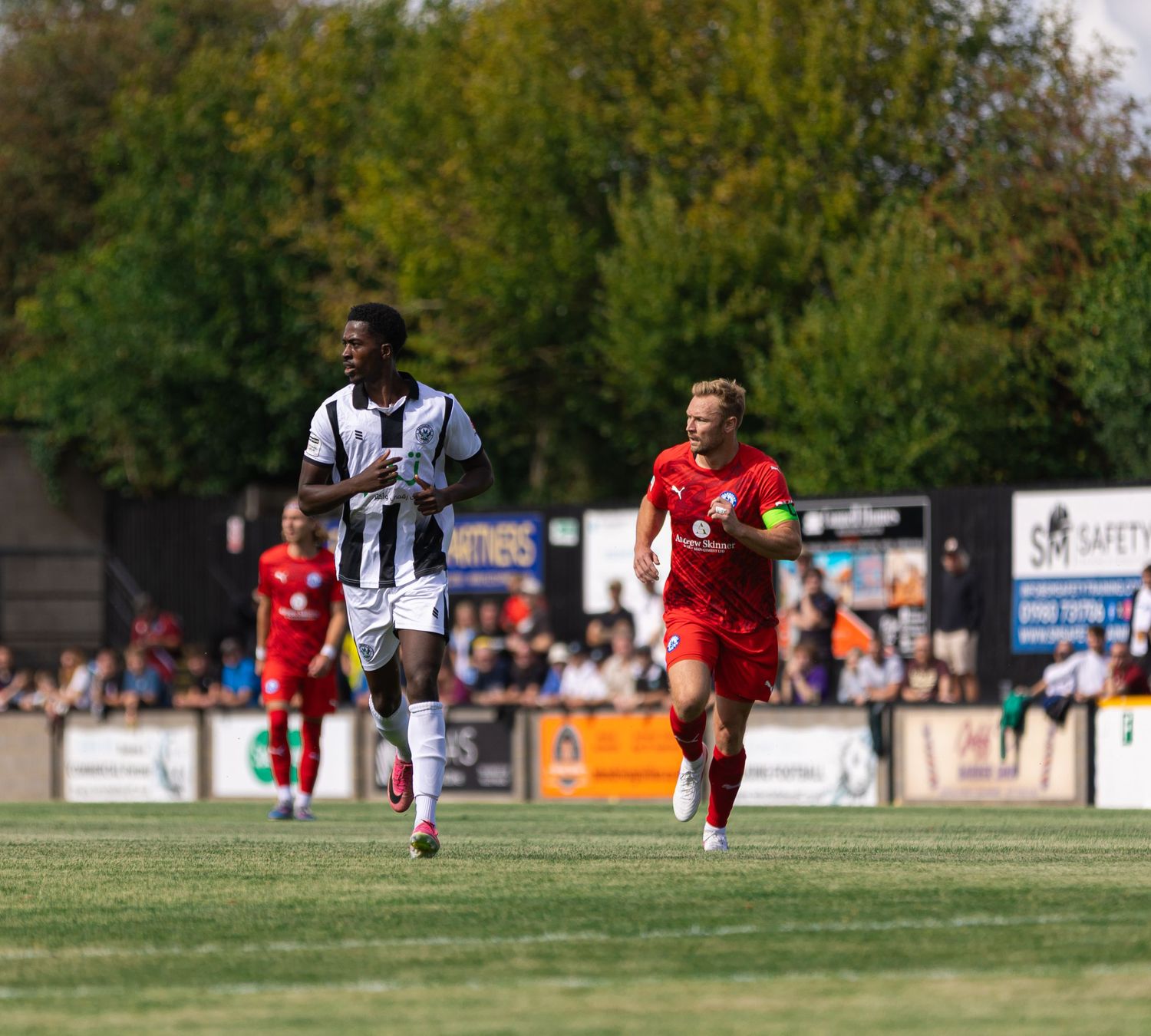 Two football players running on a grass pitch during a sunny outdoor match, one in black and white, one in red.