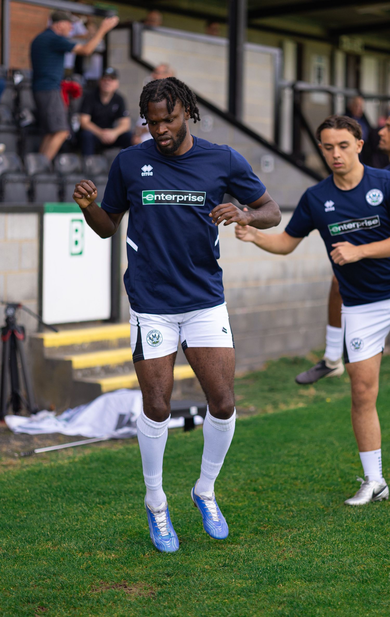 Two soccer players in navy Enterprise-sponsored Errea kits warm up on a grass pitch before a match.