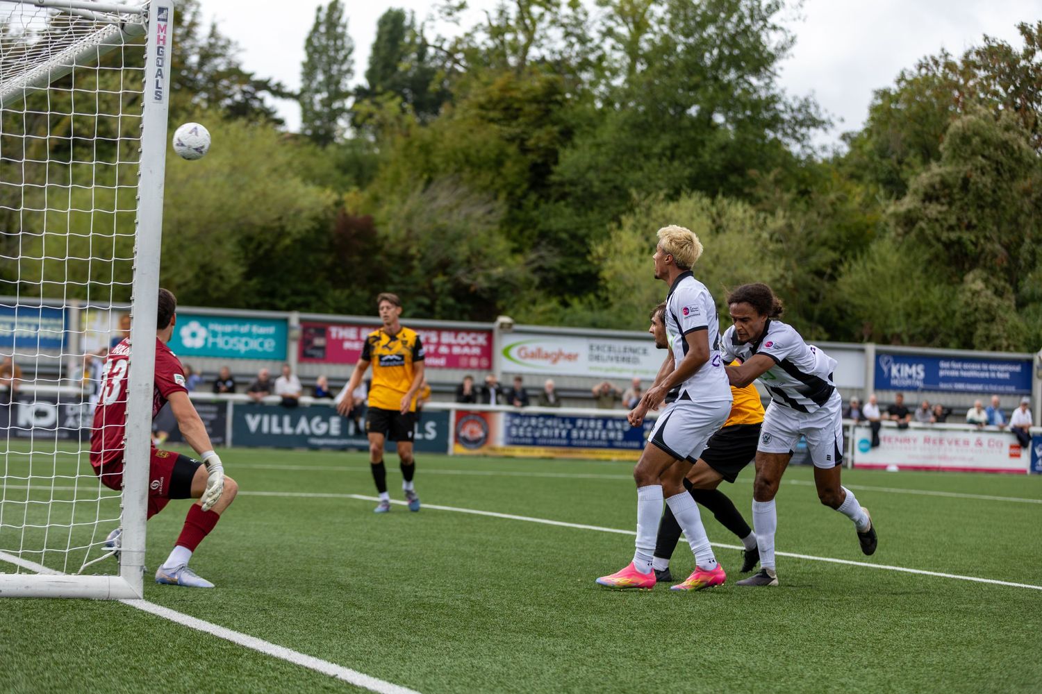 Soccer players compete near the goal during an outdoor match, with spectators and green trees visible in the background.
