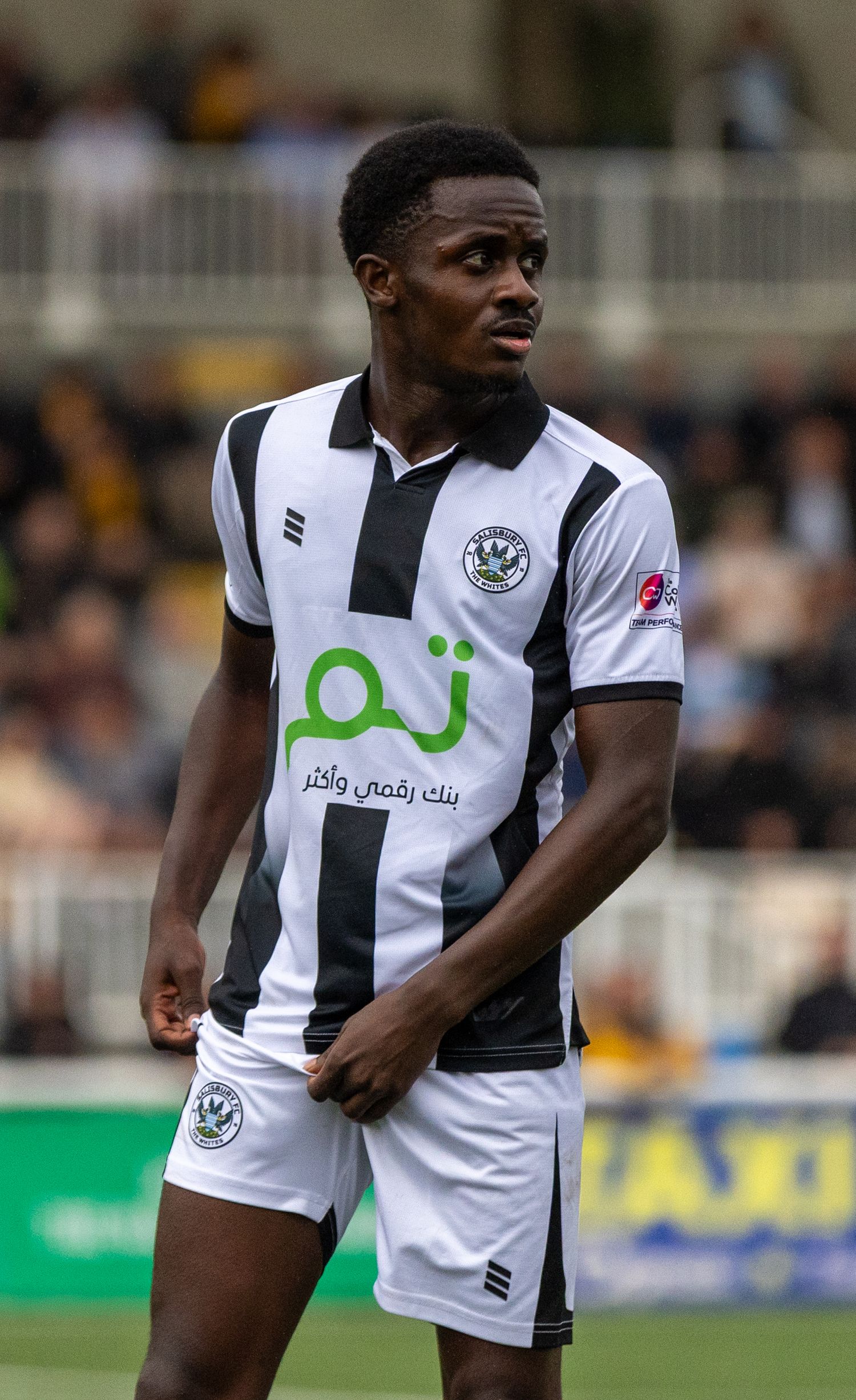 Football player in black and white striped kit with Arabic sponsor logo stands on pitch during match.