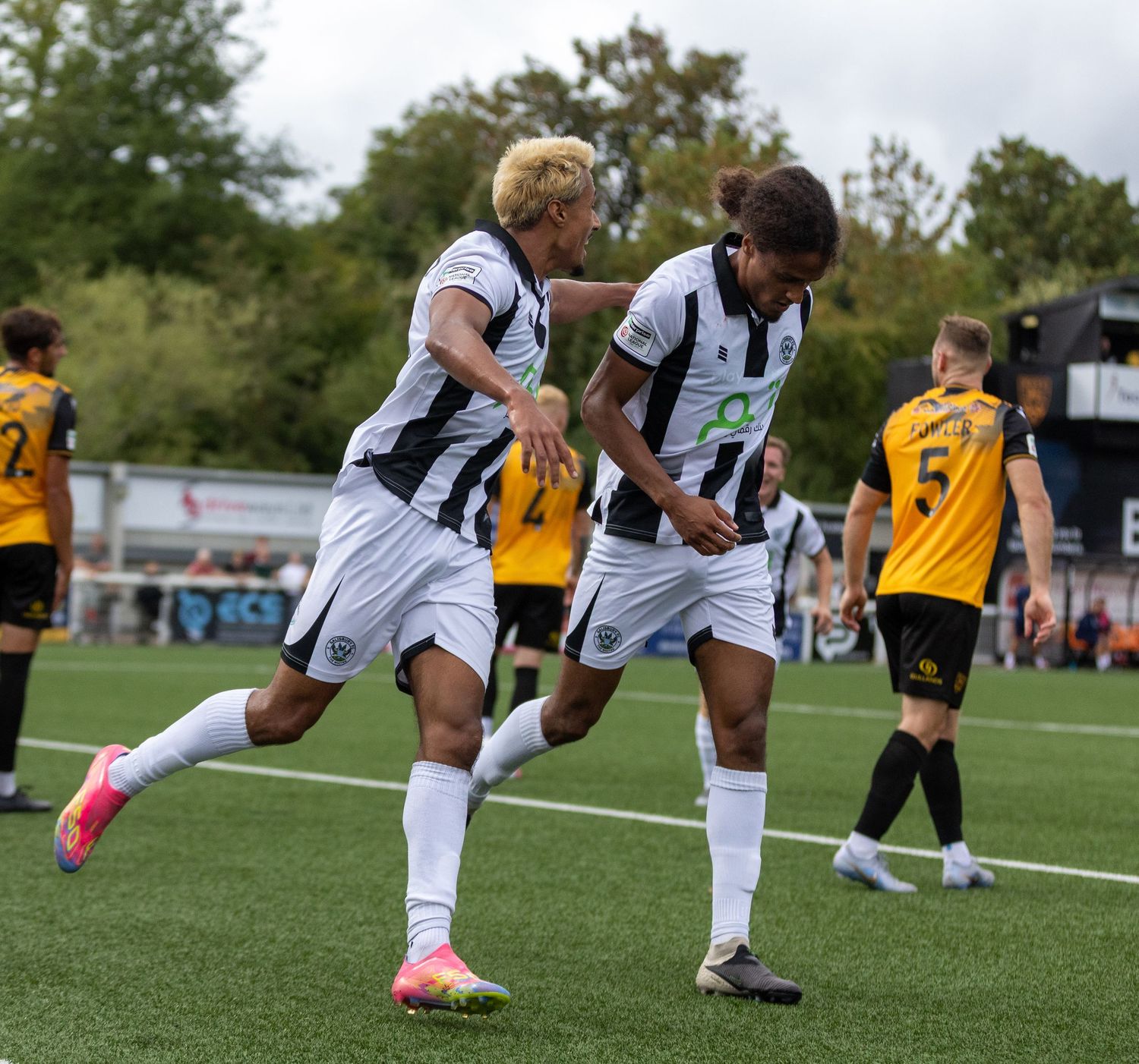 Two soccer players in black and white uniforms celebrate a goal during a match, with opponents in yellow visible in the background.