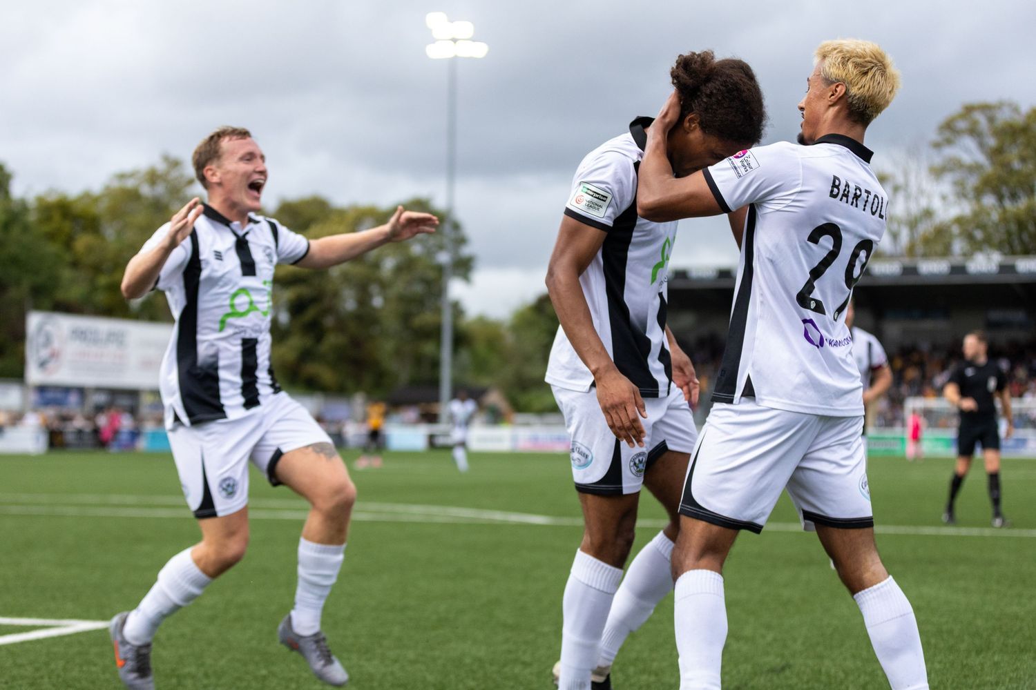 Soccer players in black and white jerseys celebrate a goal together on a sunny outdoor pitch during a match.