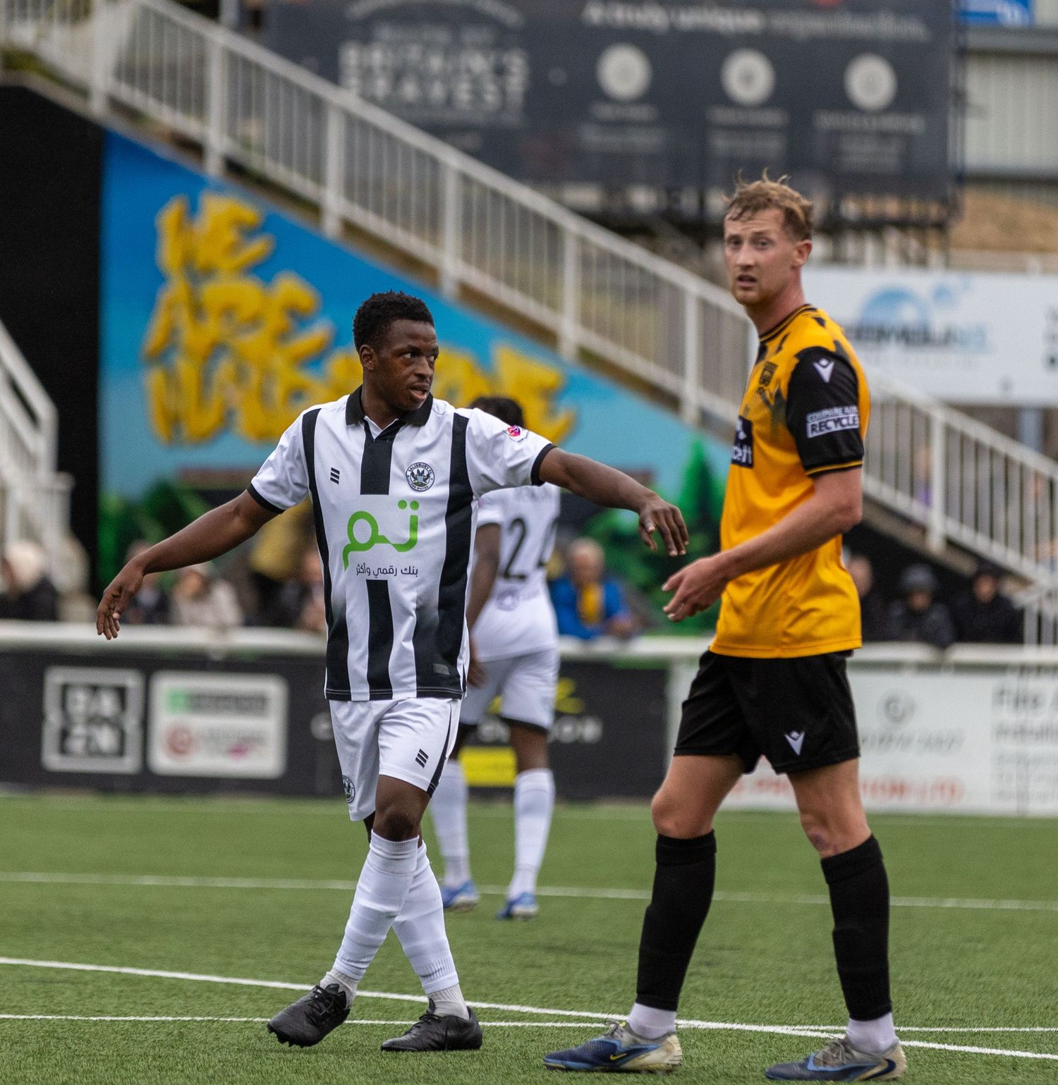 Two soccer players in contrasting black-white and yellow jerseys compete on a green football pitch during a match.