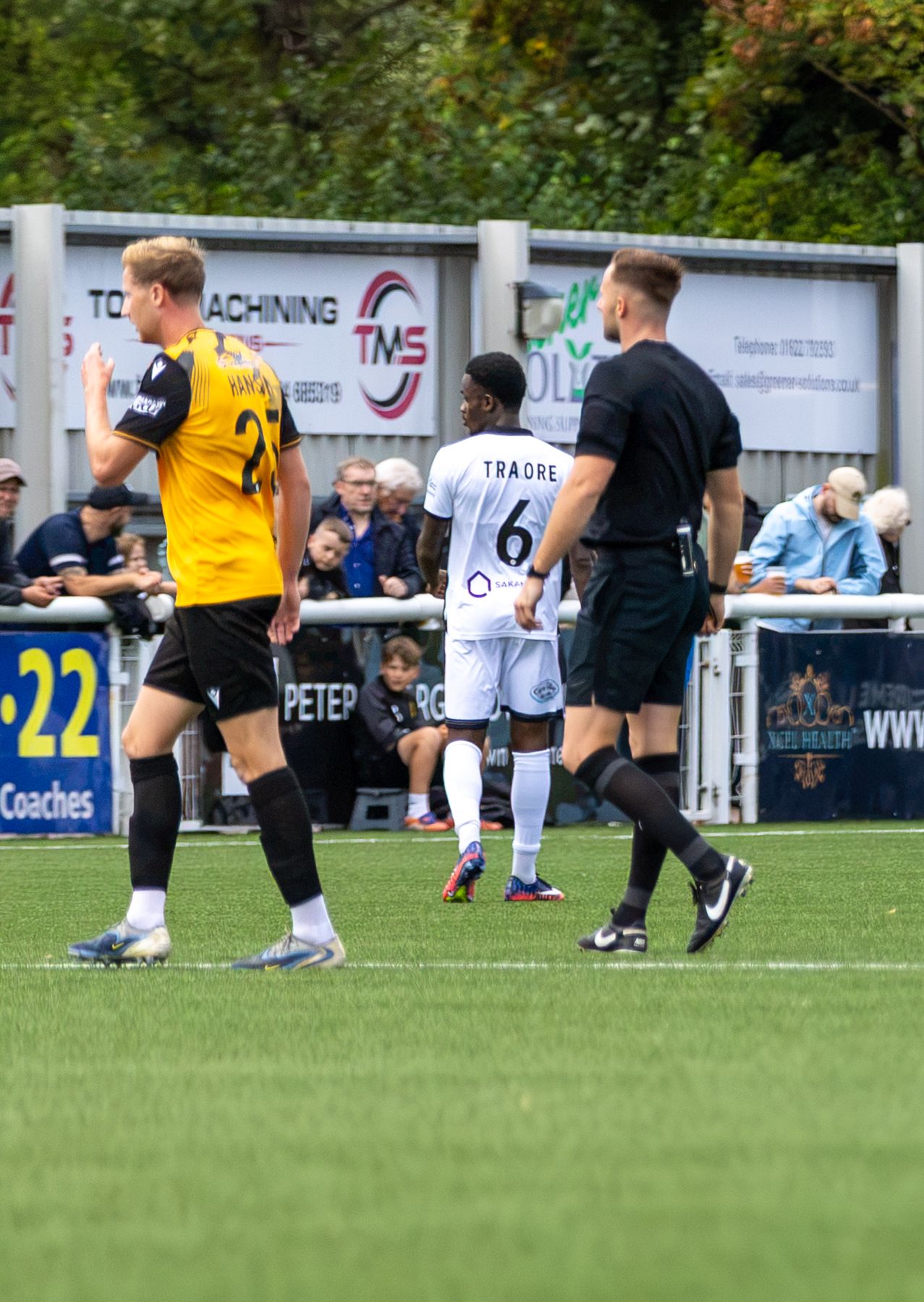 Football players including Traore number 6 in white and a player in yellow-black kit on a grass pitch with spectators.