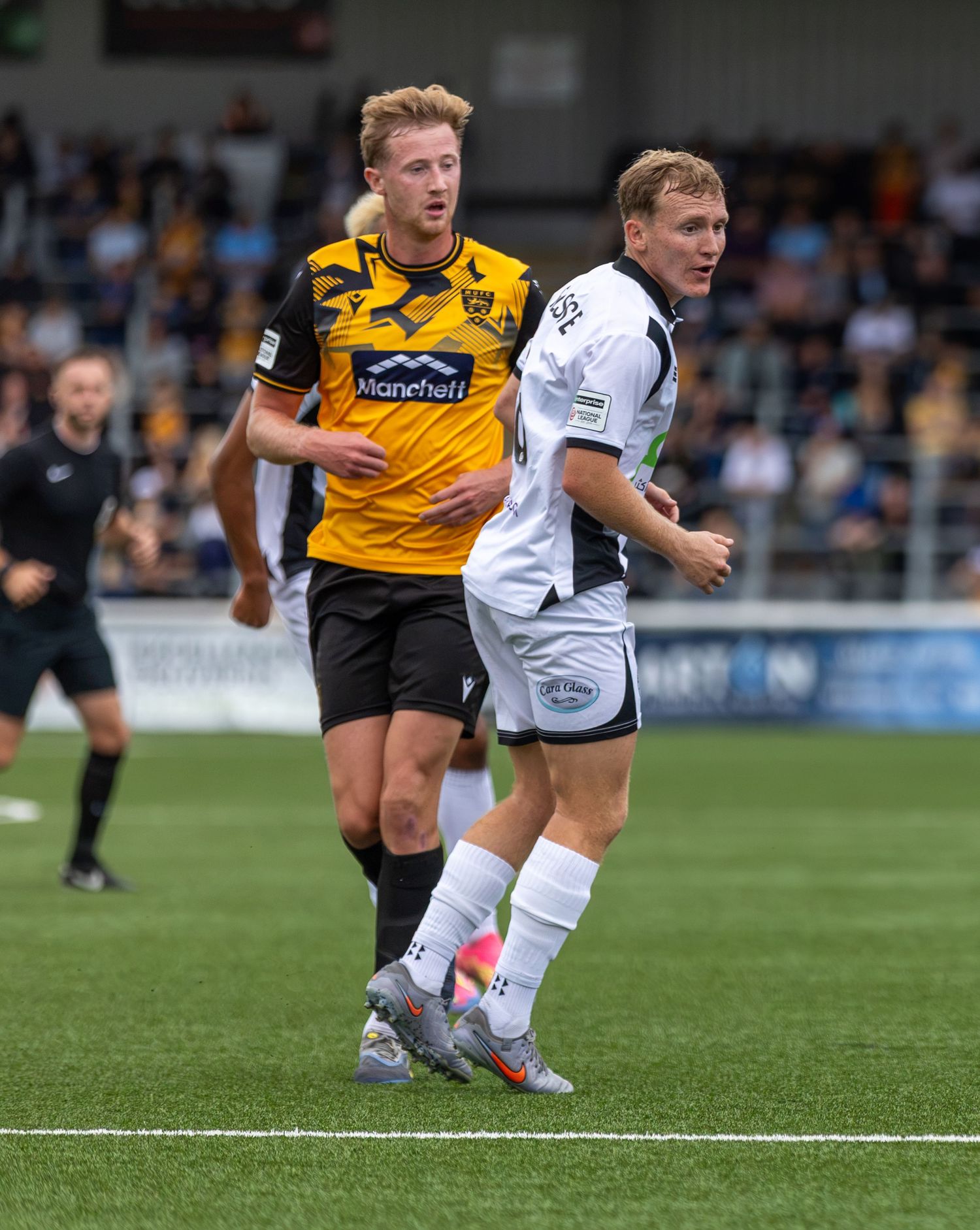 Two soccer players compete on a grass field, one in black and yellow, the other in white, during a match.