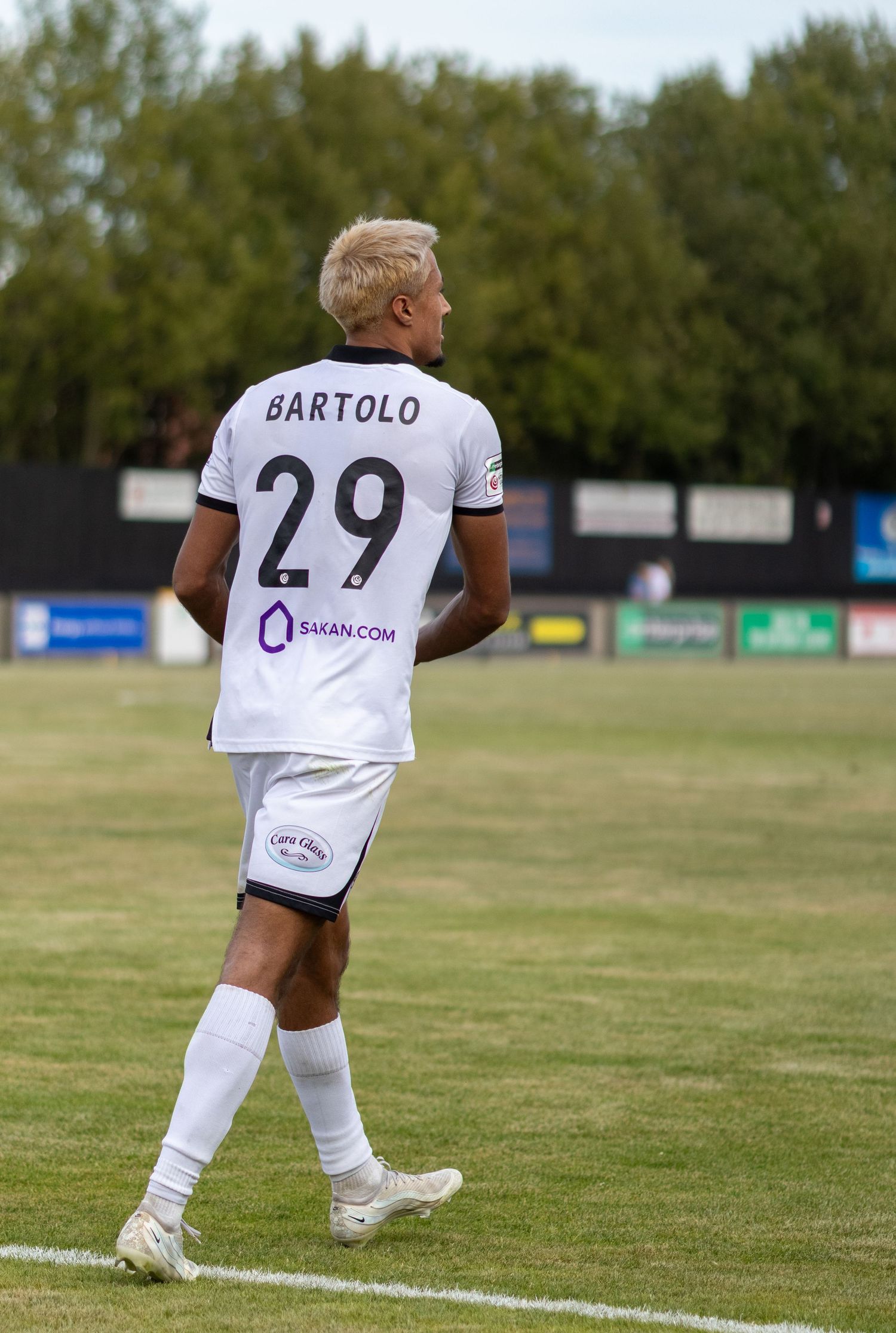 Footballer Bartolo wearing number 29 white jersey with SAKAN.COM sponsor walks on pitch.