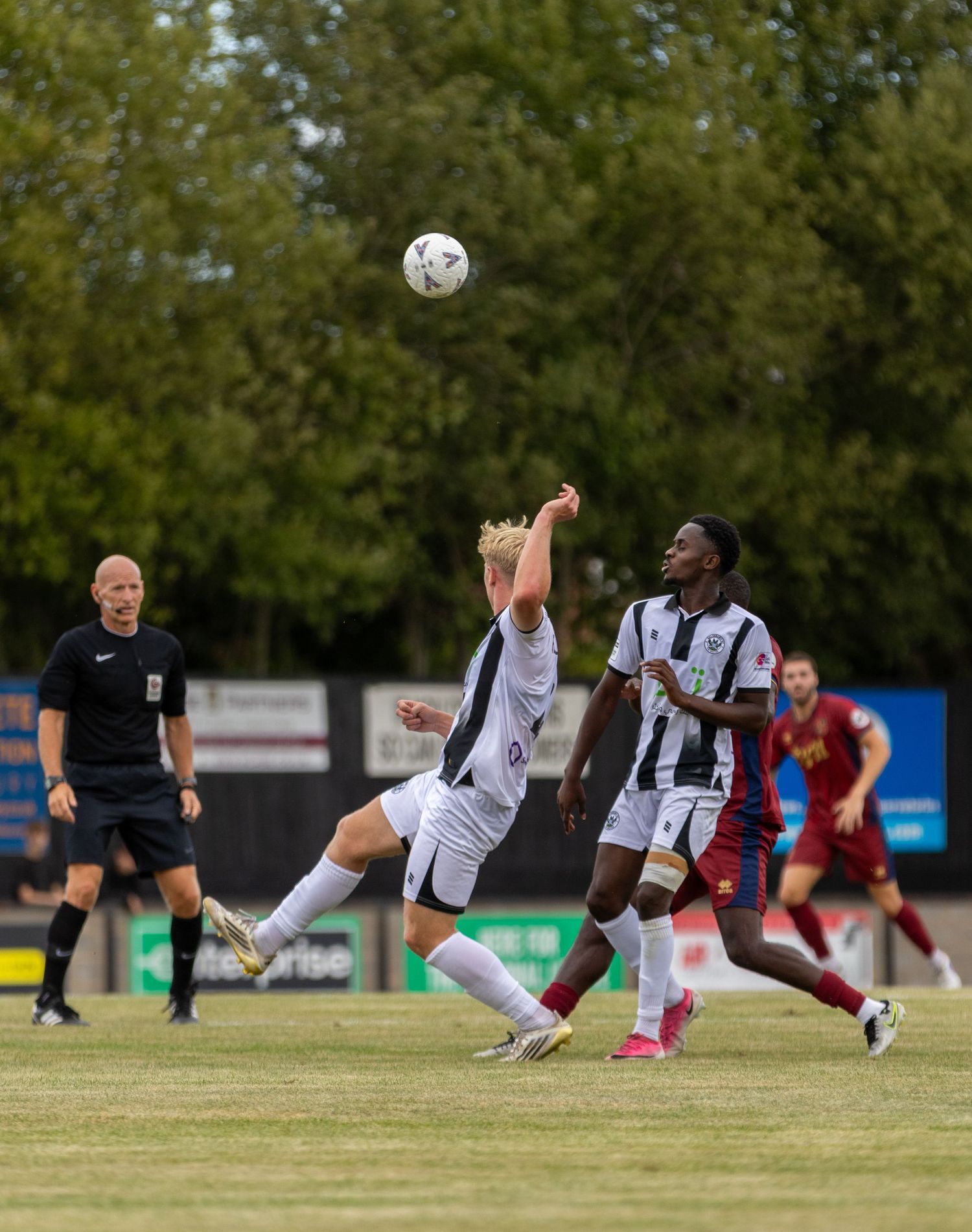 A referee watches as two footballers compete for a high ball during a non-league match on a sunny day.