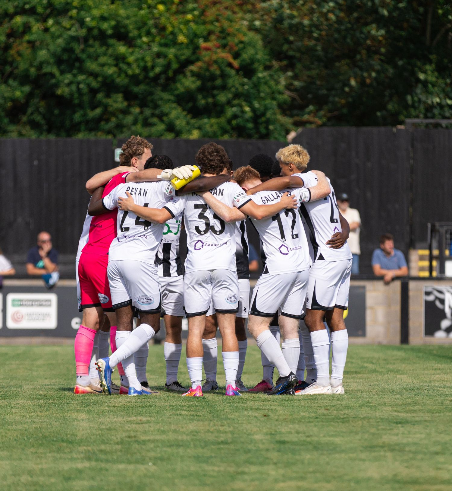 Soccer team huddles together on sunny field, wearing white uniforms with numbers, preparing for match.