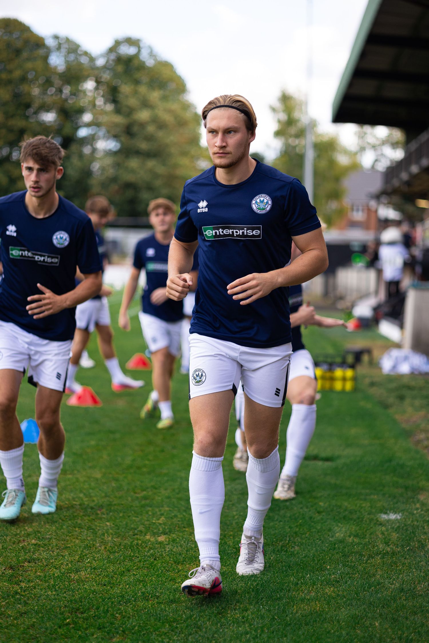 Football players in navy Enterprise-sponsored kits warming up on a green pitch during pre-match training session.