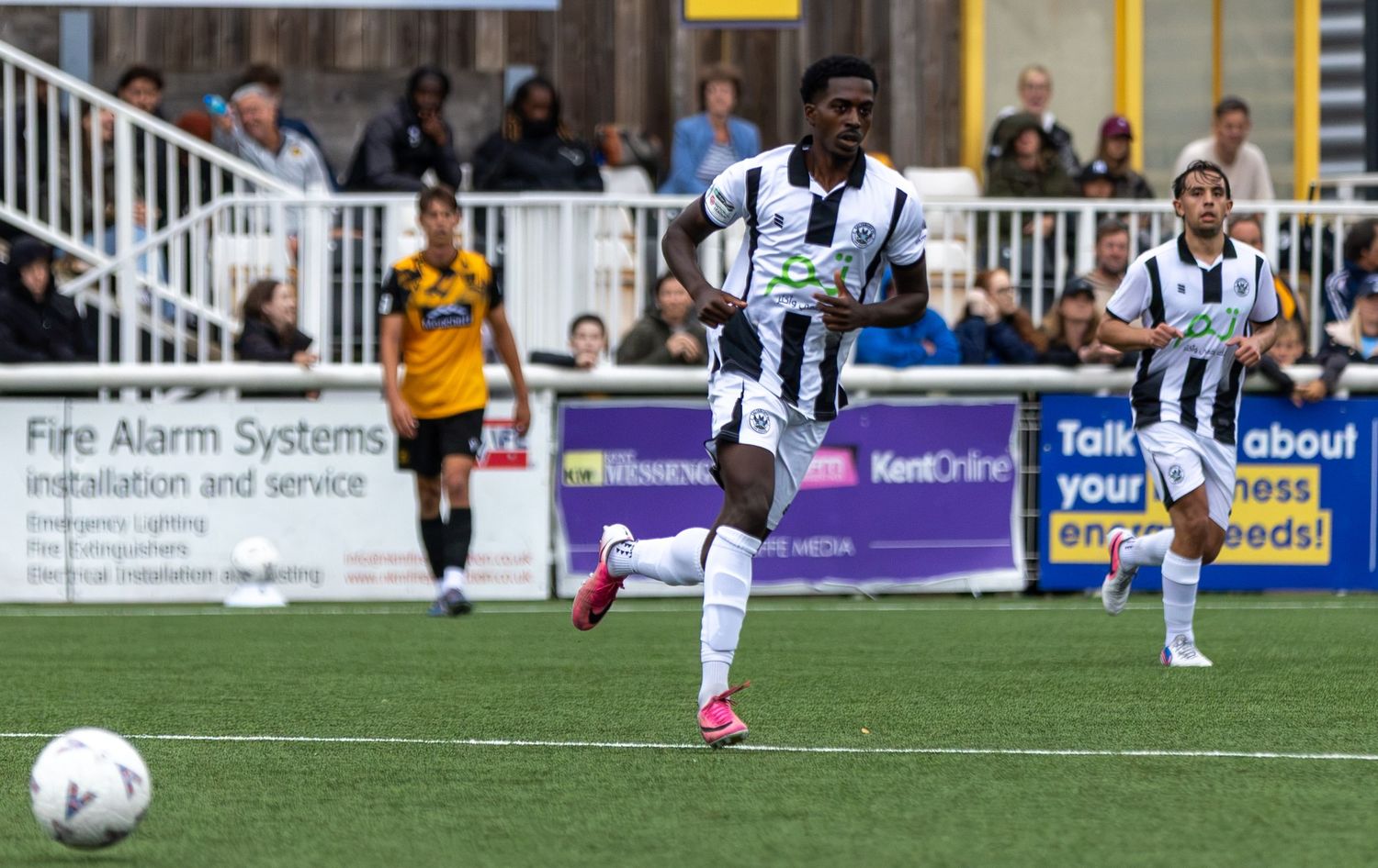 Soccer player in black and white striped kit dribbles ball during a match at a stadium.