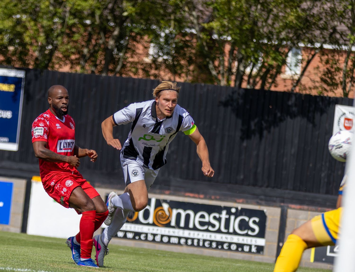 Soccer players in red and black-white kits sprint during a competitive football match on a sunny day.