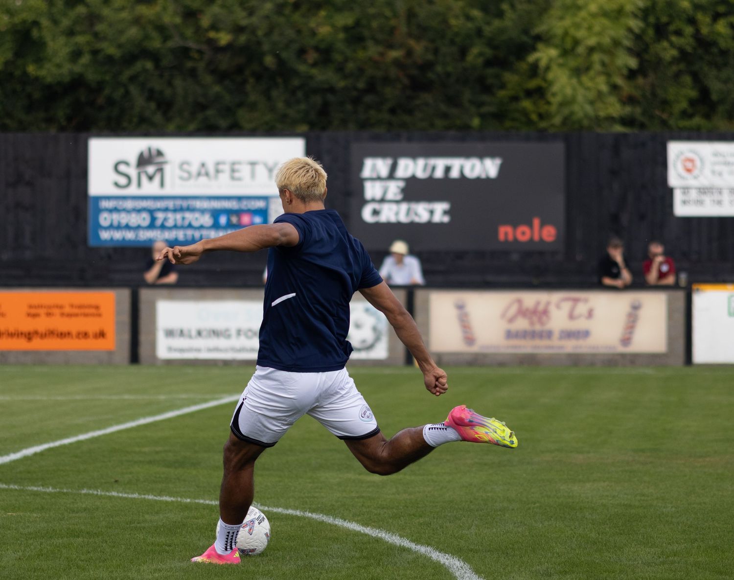 Footballer in navy blue kit taking a shot during pre-match warmup with In Dutton We Crust banner visible.