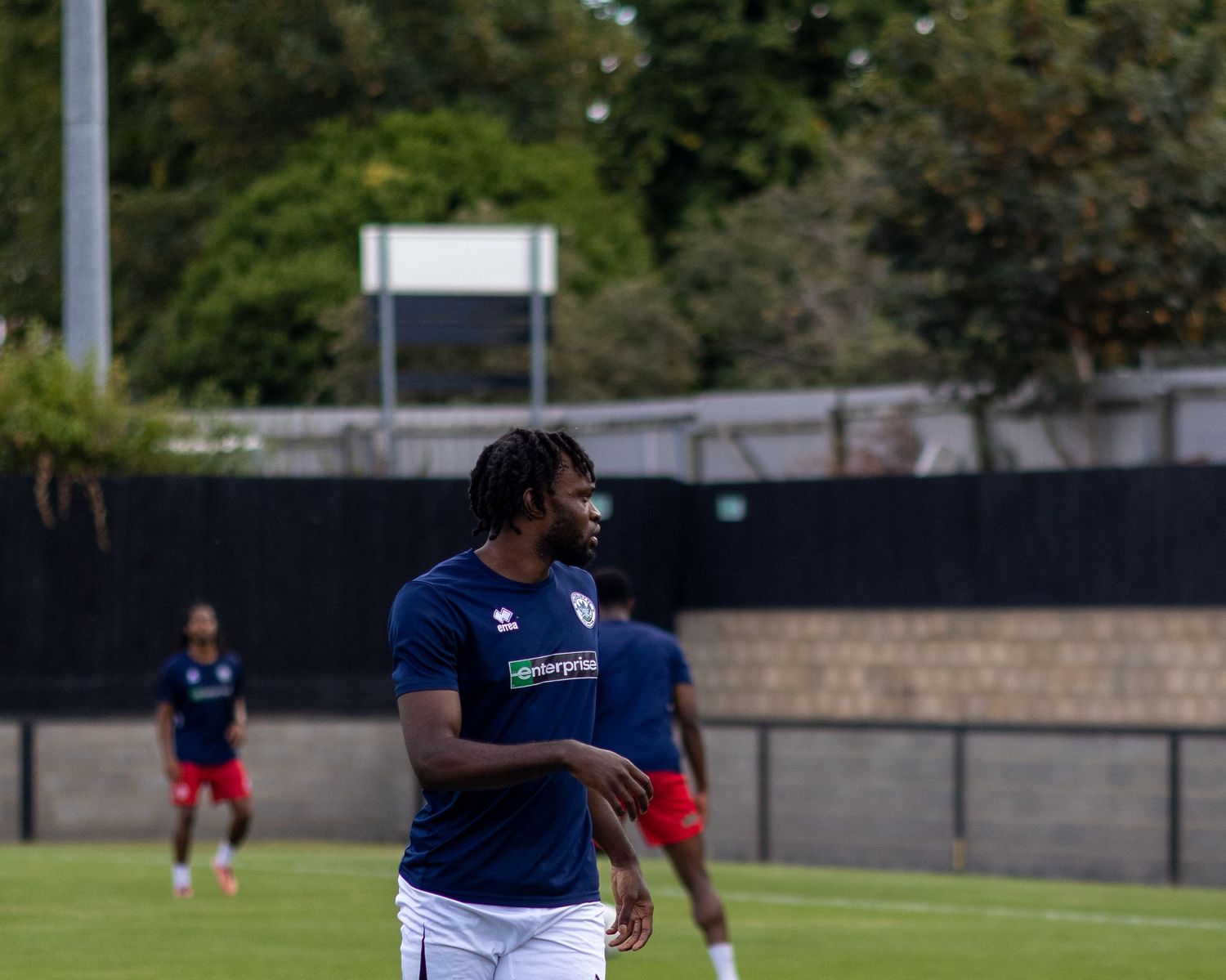 Male footballer in navy enterprise-sponsored kit jogs across a grass pitch during a pre-match warmup session.