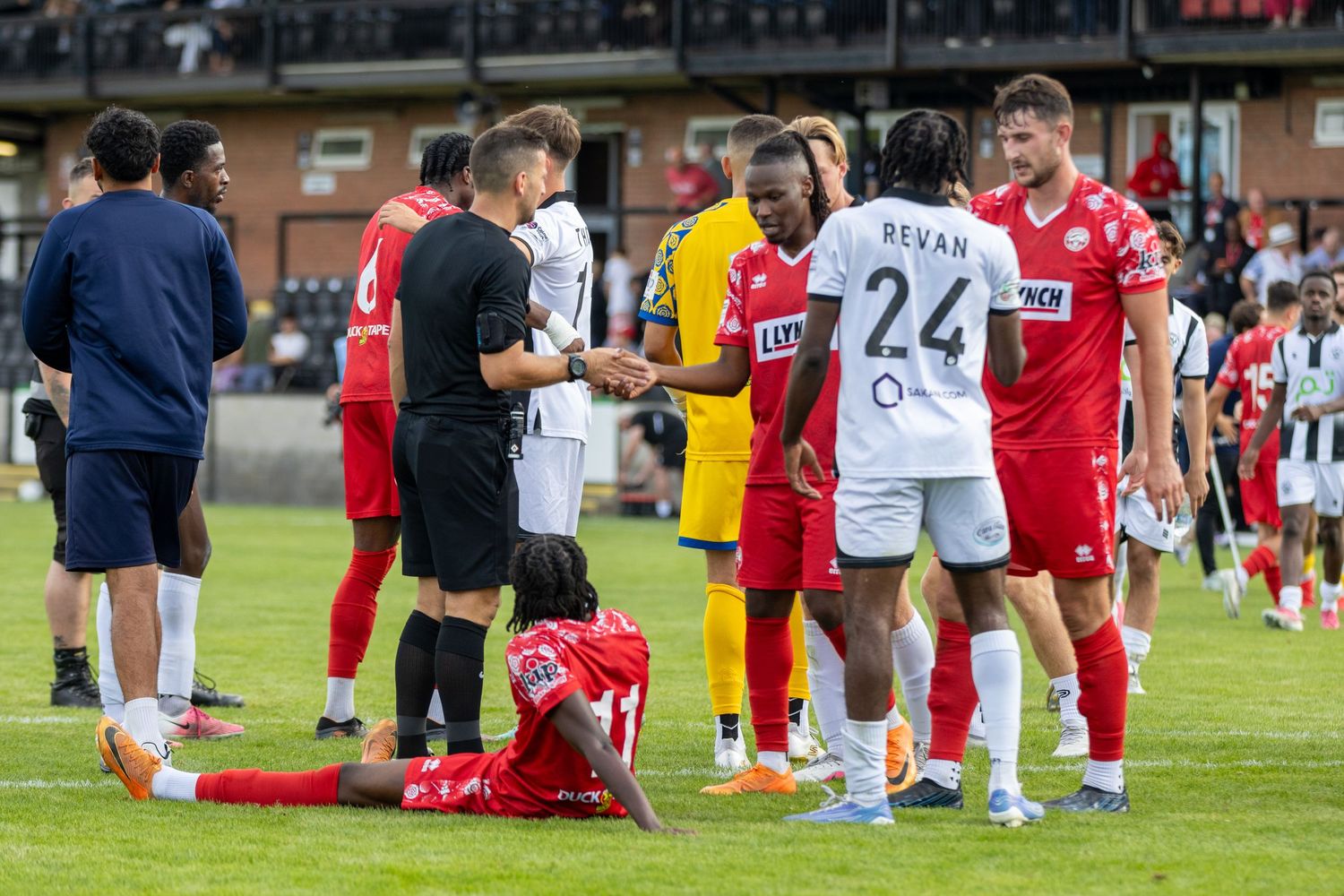 Soccer players and referee gather on field during match, with player sitting on ground and number 24 jersey visible.