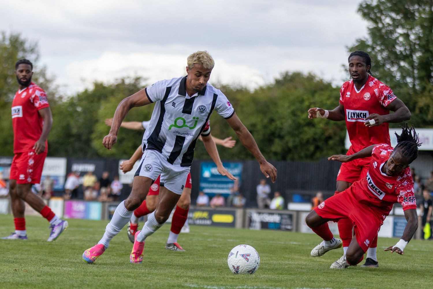 A footballer in black and white kit charges forward with the ball past sliding opponents.