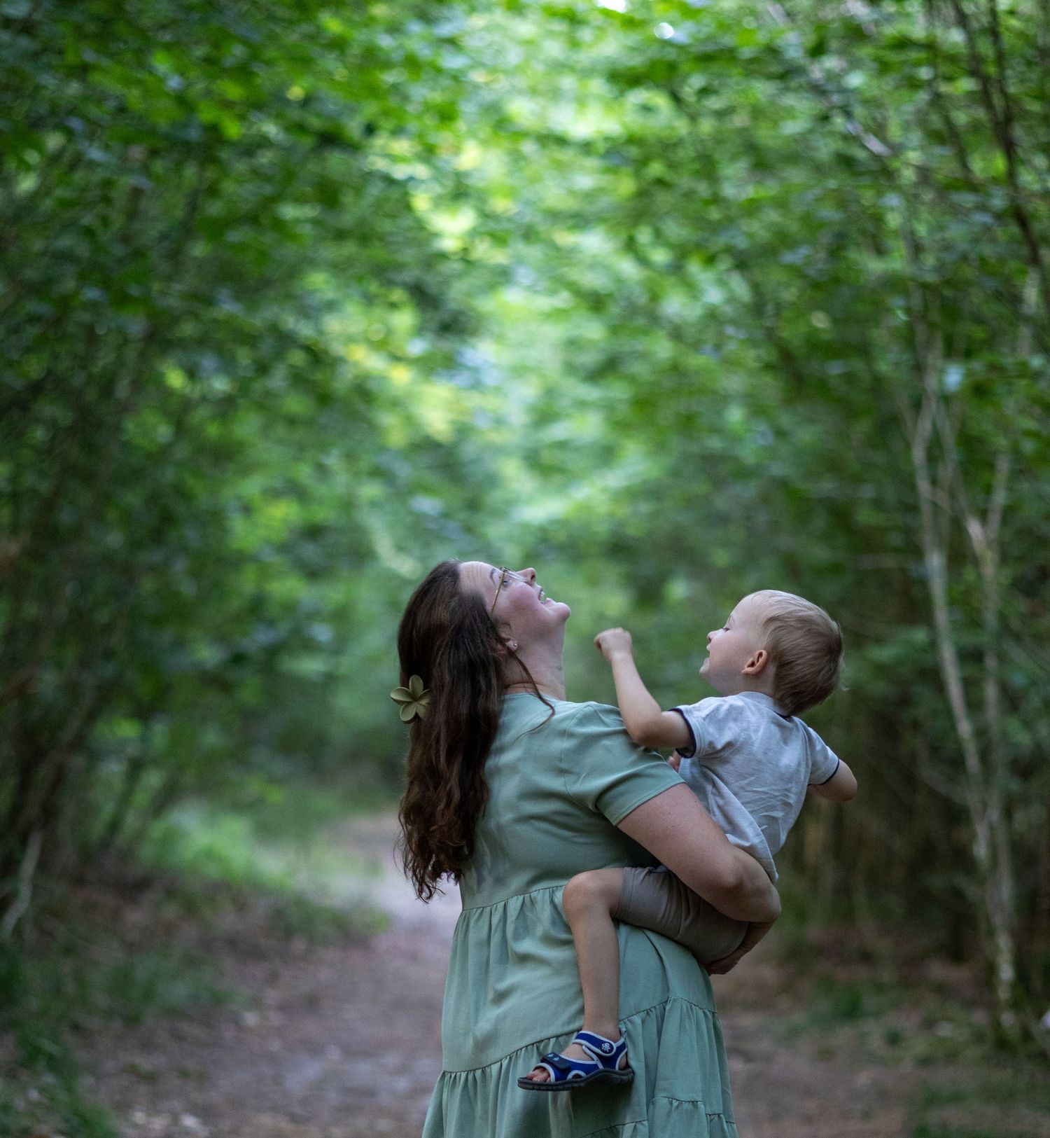 Mother in mint dress holding baby boy on woodland path, looking up joyfully.