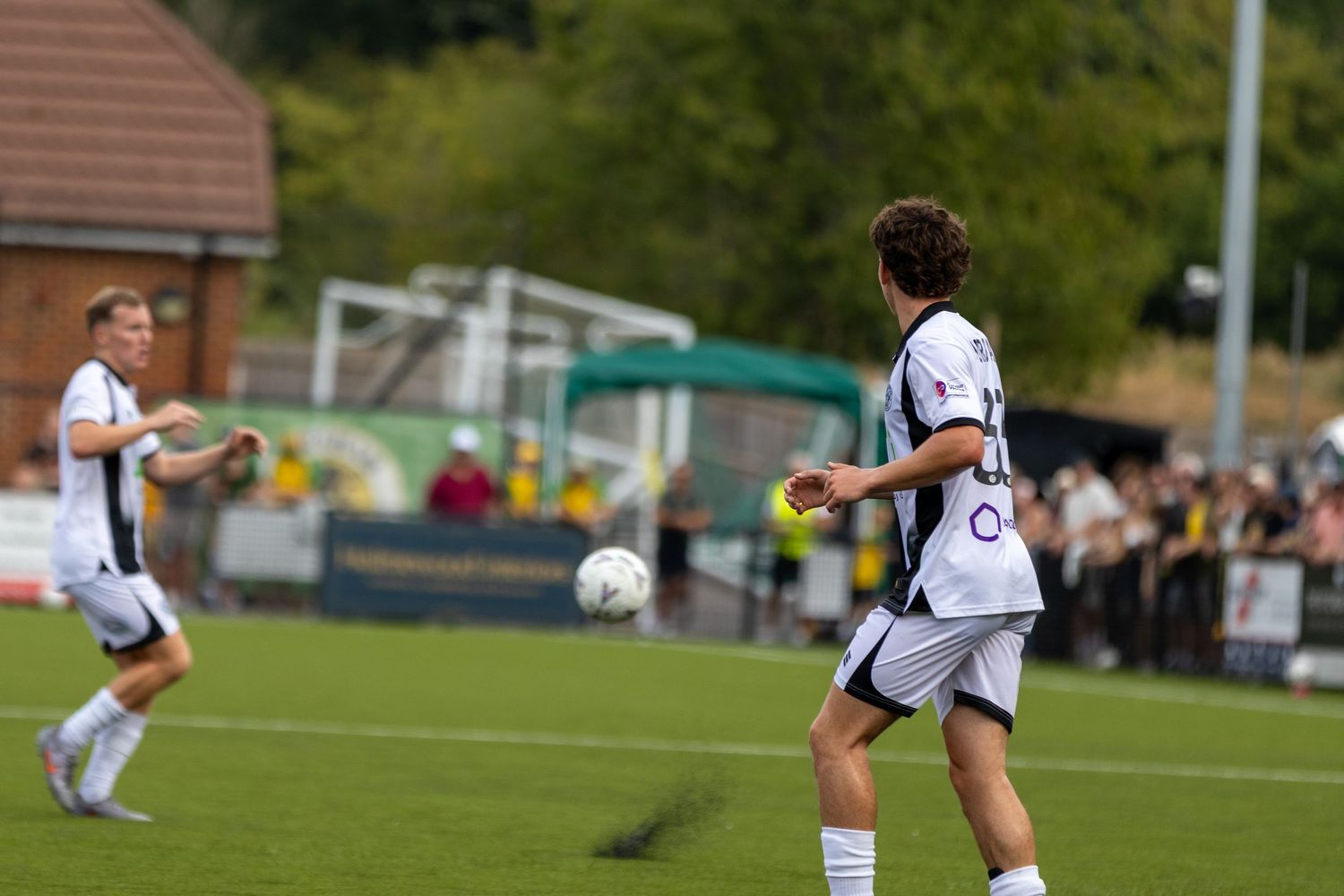 Two footballers in white kits pass the ball during an outdoor match on a green pitch.