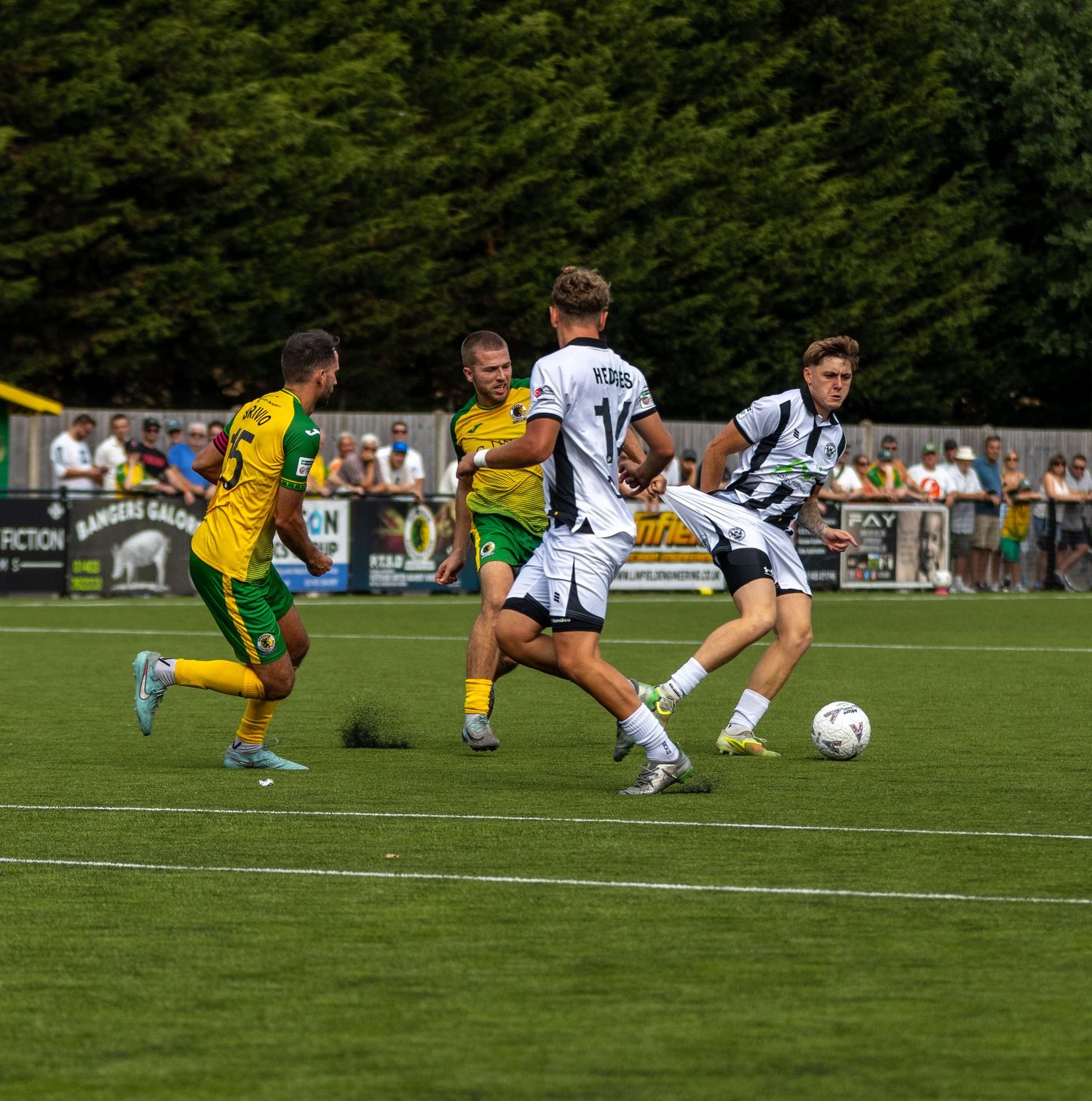 White-kitted footballer dribbles past yellow-green opponents on a grass pitch during a sunny non-league football match.