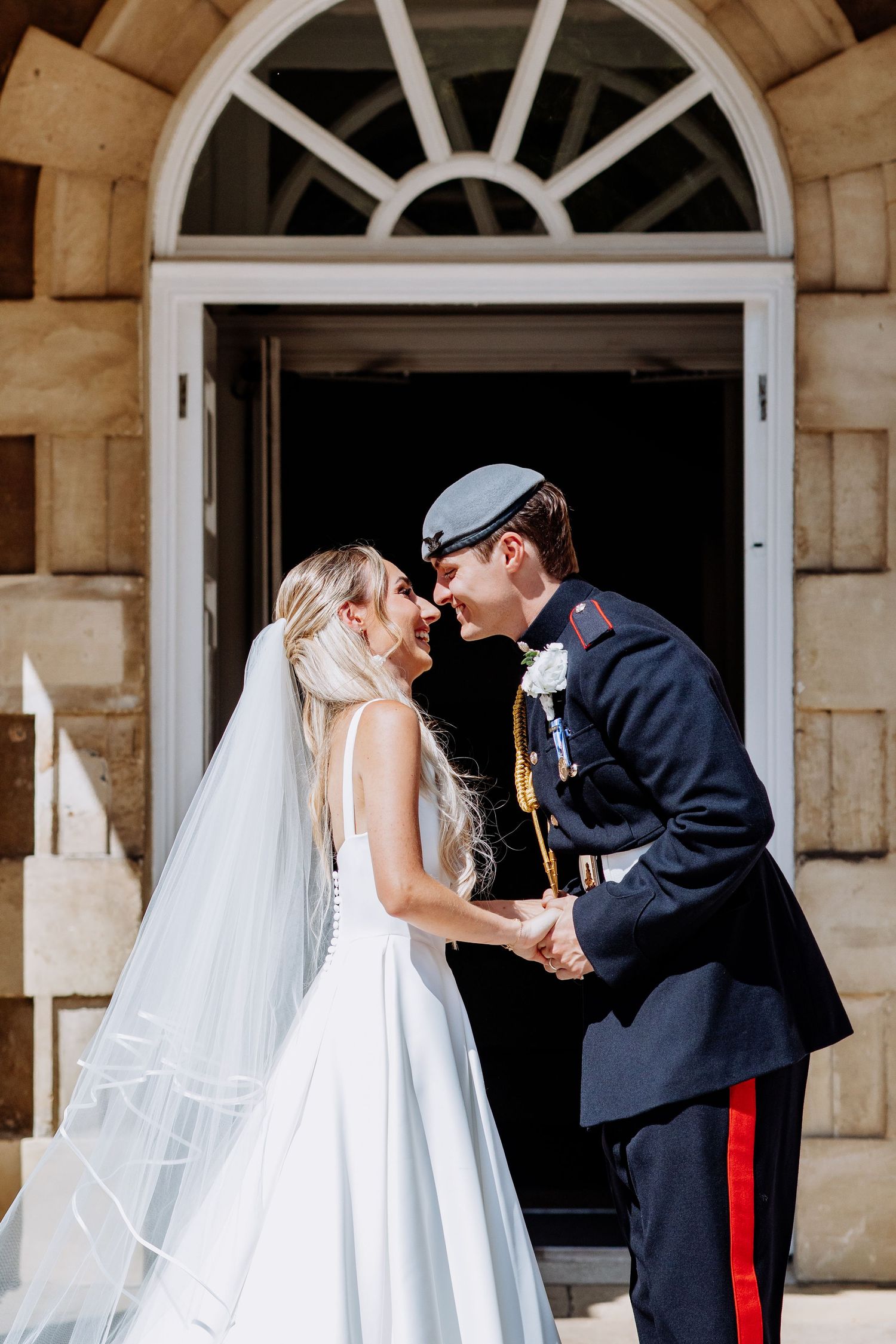 Bride in white gown and veil holds hands with groom in military uniform as they lean in to kiss.