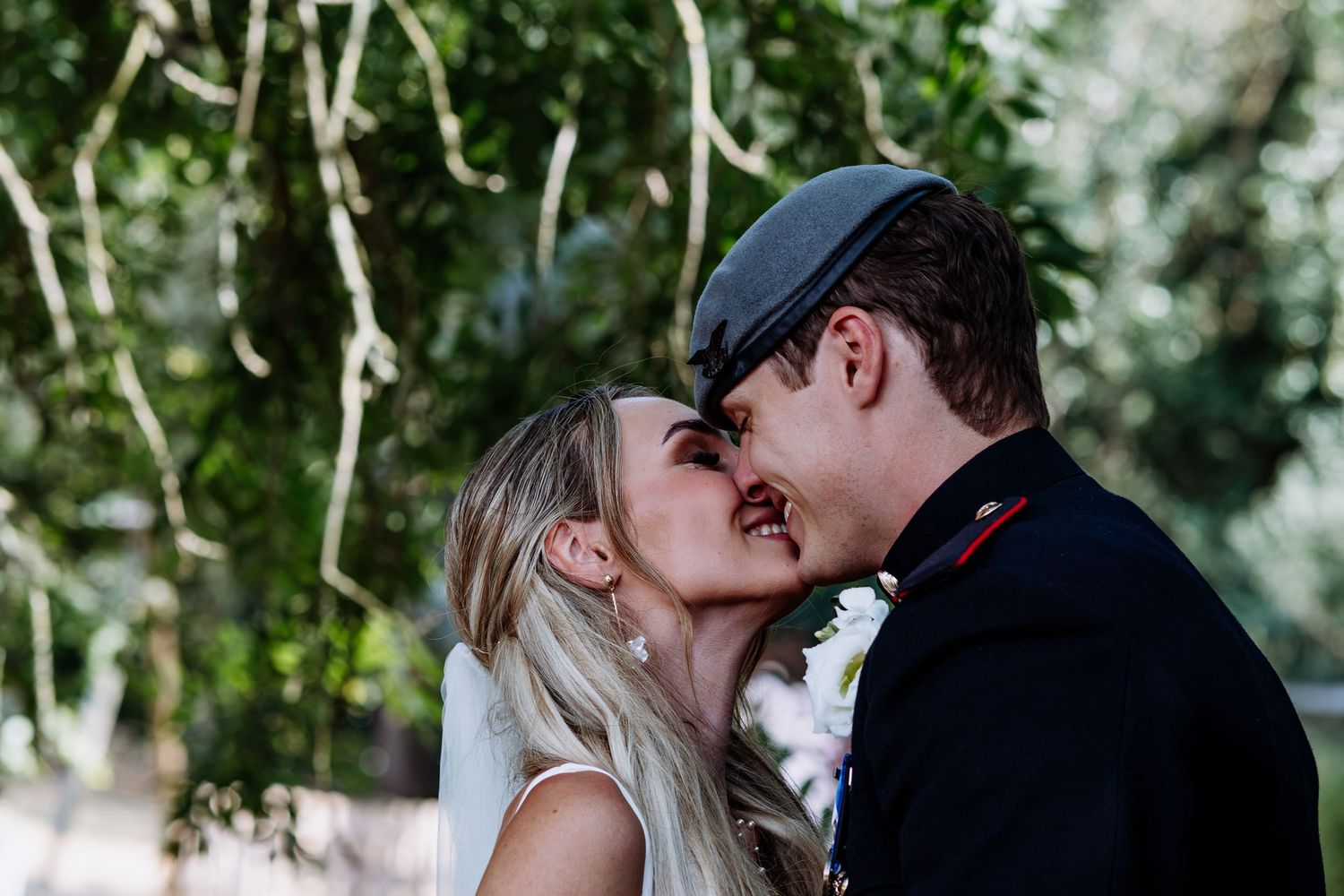 Bride and groom kiss tenderly surrounded by lush green trees outdoors.