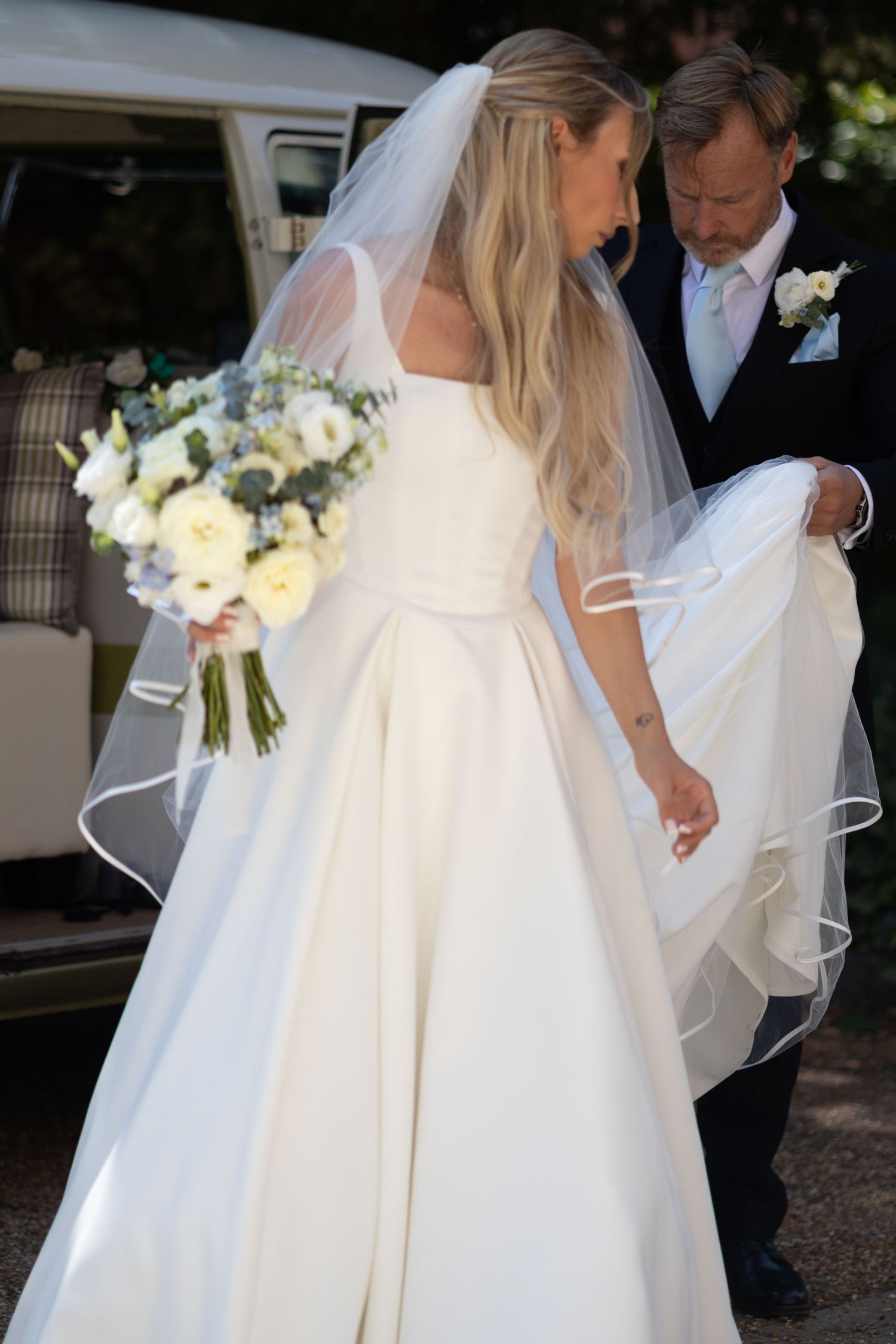 A bride in a full white ball gown holding a bouquet stands with her father beside a vintage VW campervan.