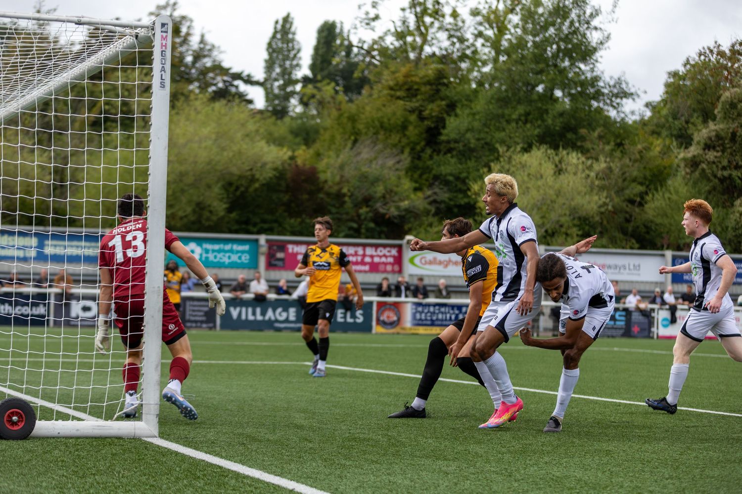 Football players compete near the goal during an outdoor match, with a goalkeeper in red and field players in black and yellow.