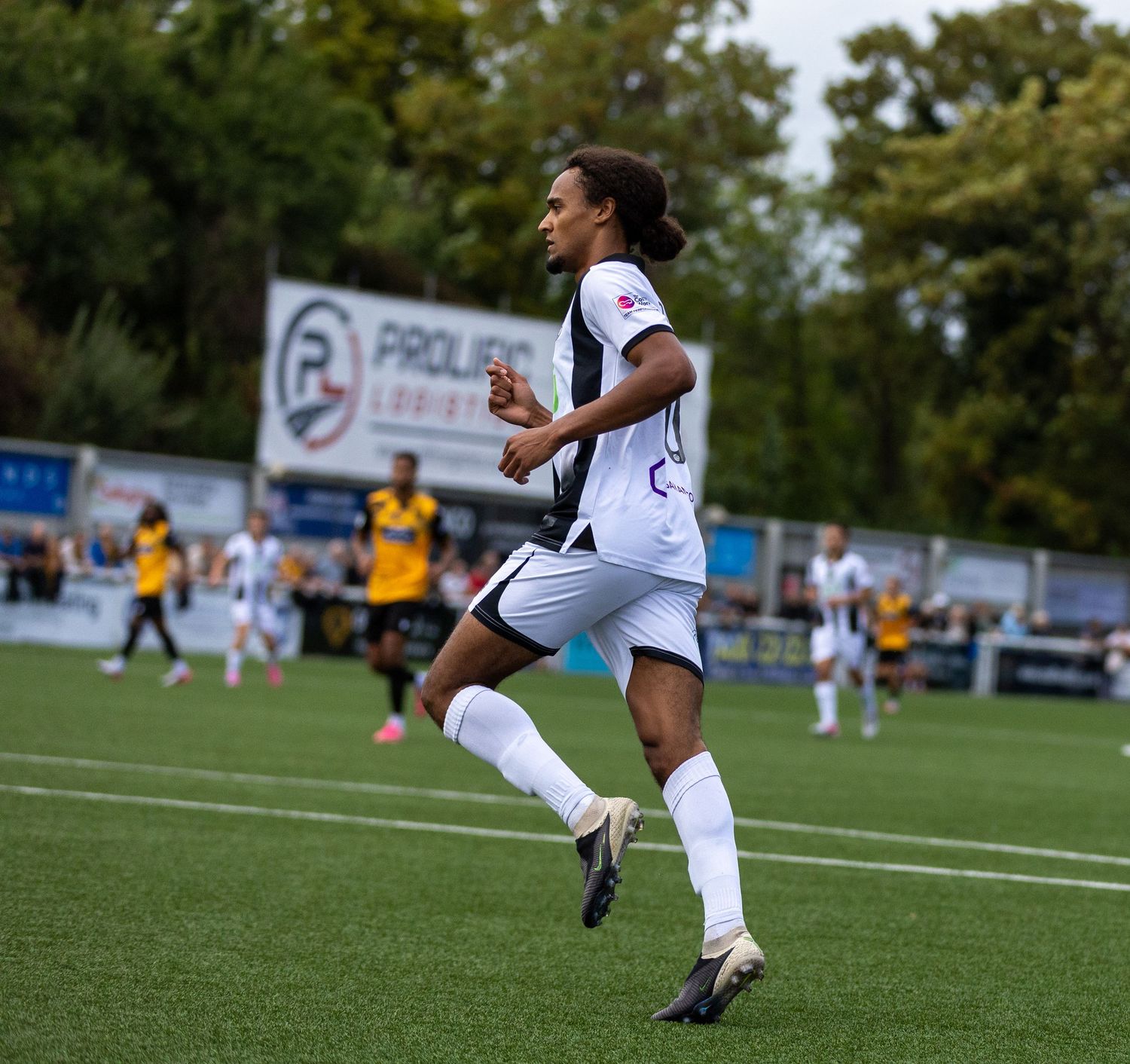 A soccer player in white uniform runs on a green field during a match with spectators and a sponsor banner in the background.