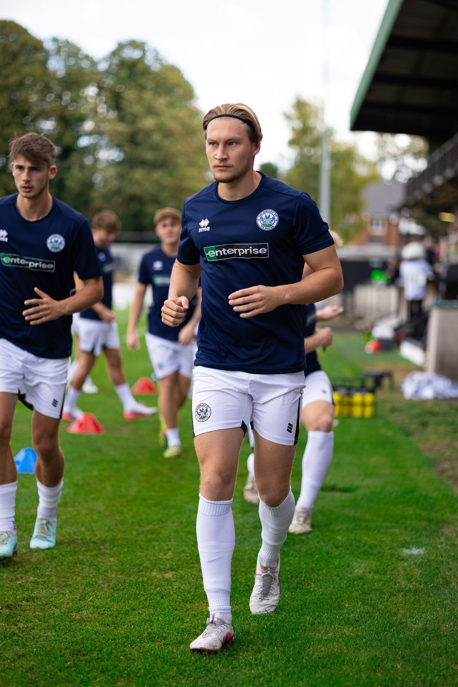 Football players in navy Enterprise-sponsored kits jogging during pre-match warm-up on a green pitch.