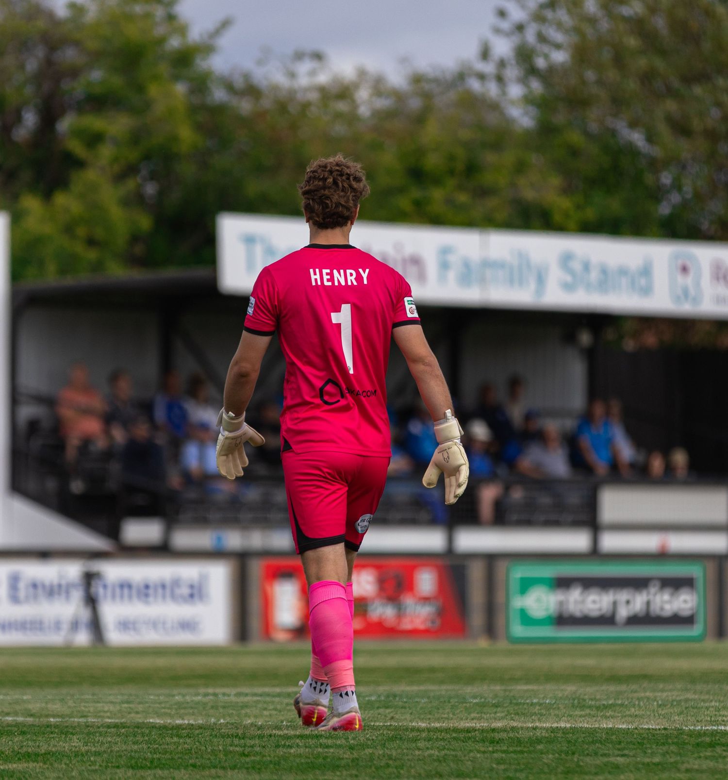 Goalkeeper Henry wearing pink kit with number 1 walks on football pitch near a stand with Enterprise and Environmental sponsors.