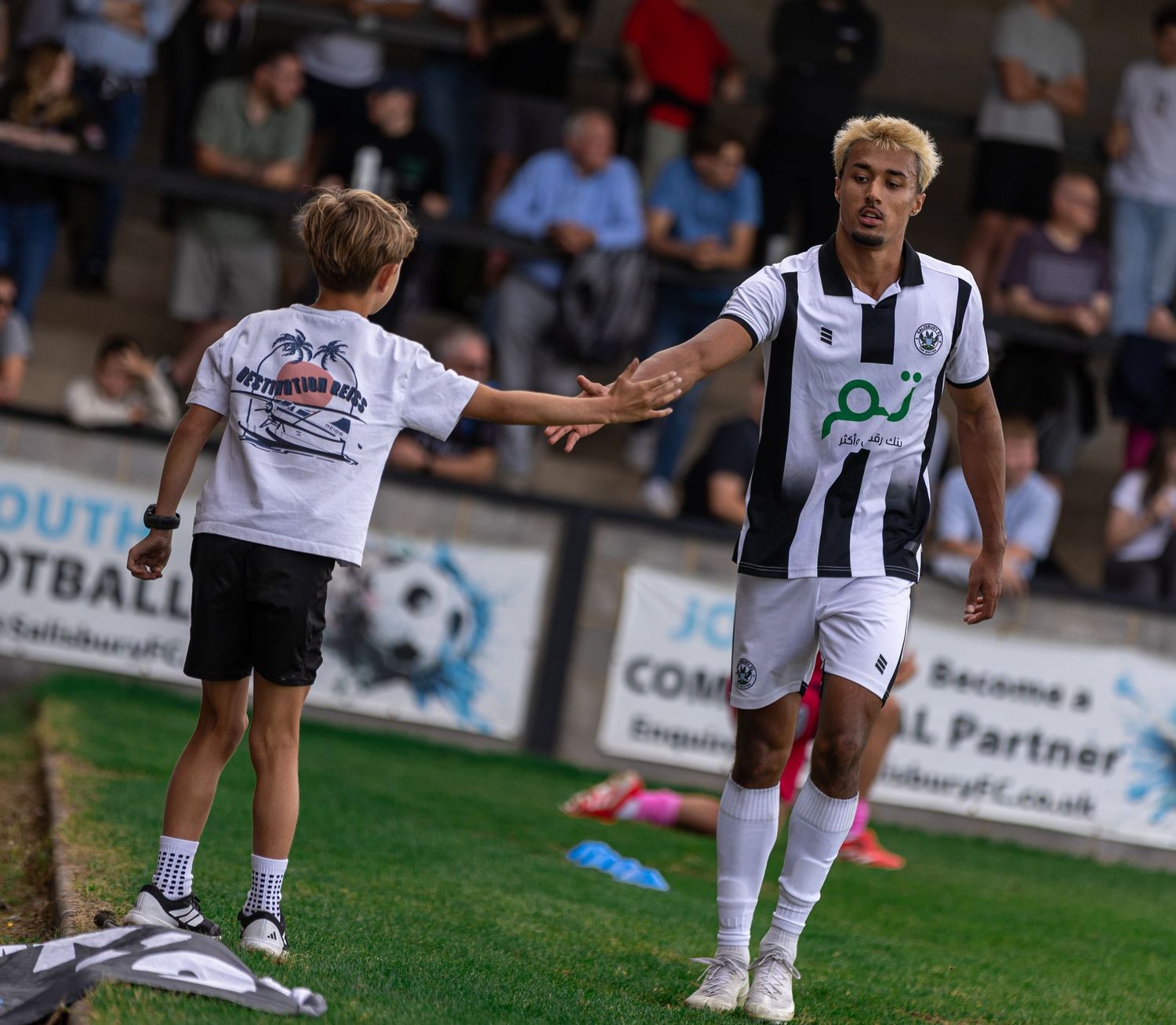 Football player in white kit high-fiving a young fan on the sideline during an evening match.