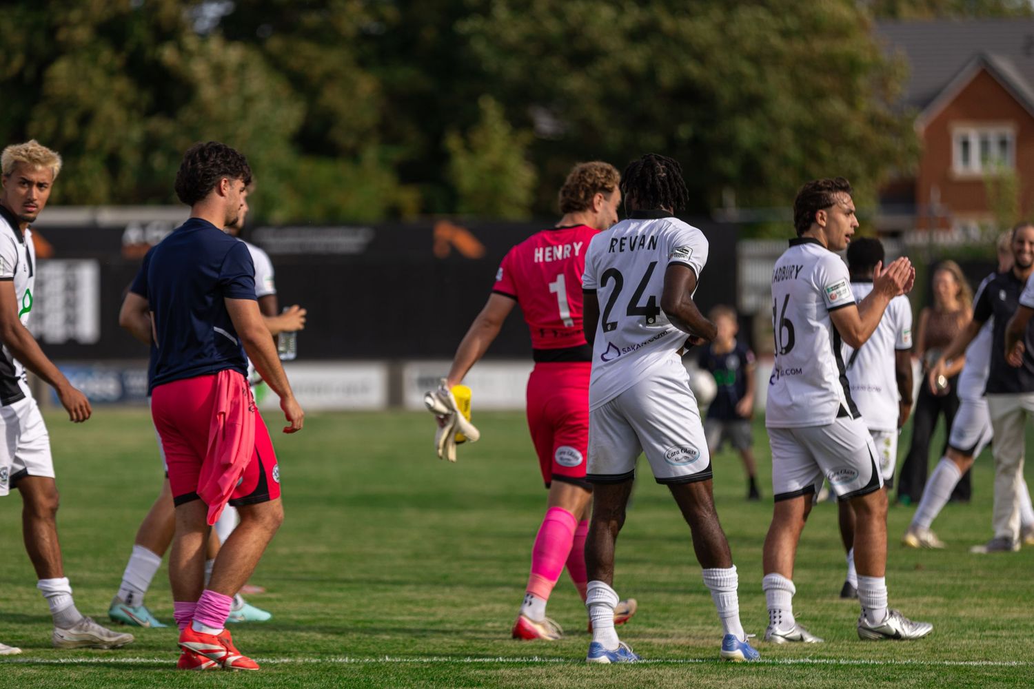 Soccer players gather on a sunny outdoor field during a match, with one player wearing a pink goalkeeper jersey.