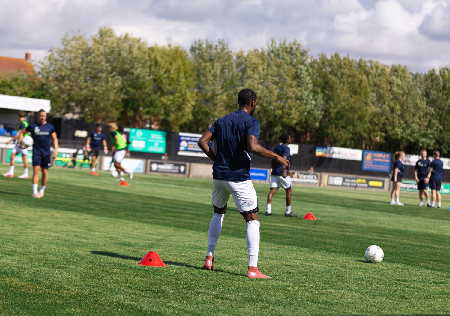 Soccer players warming up on a green pitch with orange cones during a sunny outdoor training session.