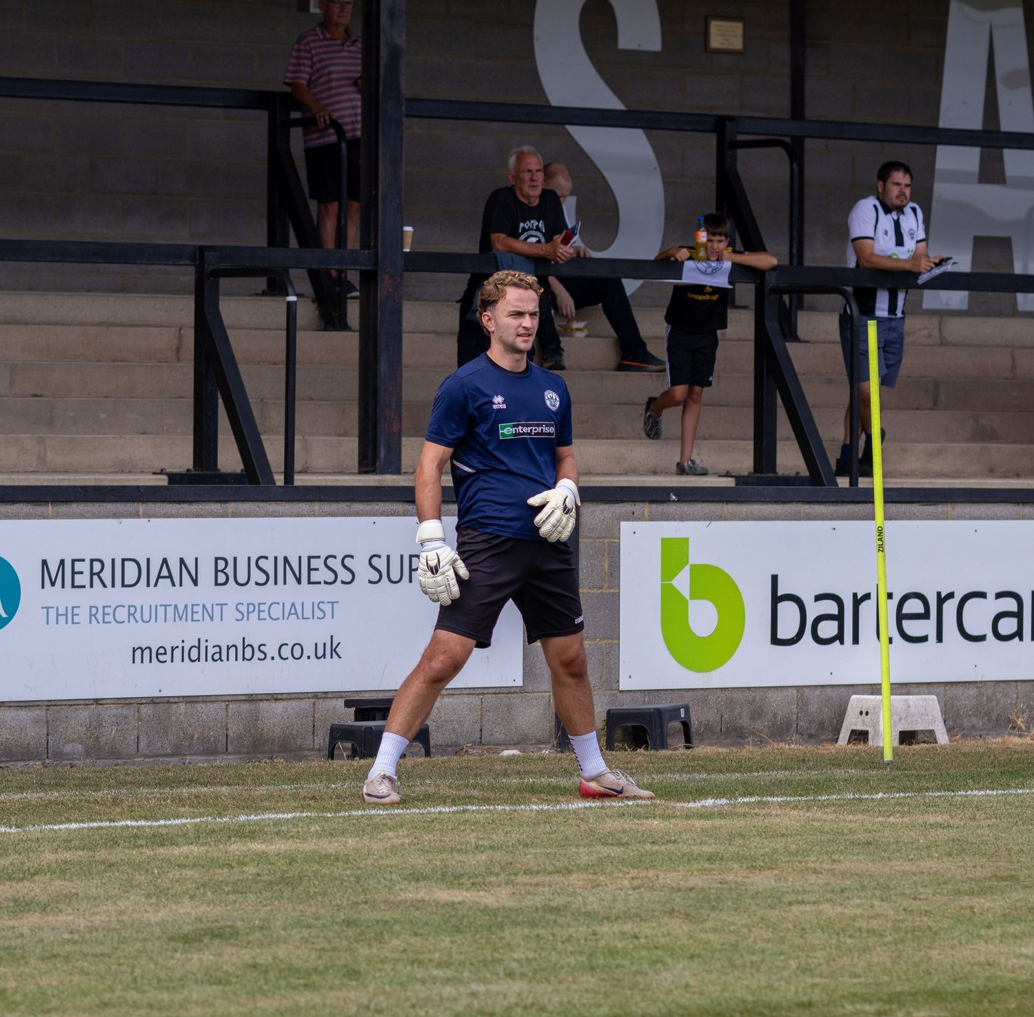 A goalkeeper in a blue kit warms up on the pitch at a non-league football ground on a sunny day.