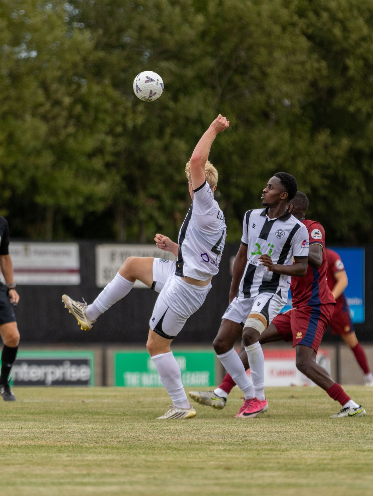 Non-league footballer stretches to reach a high ball during a competitive match on a grass pitch.